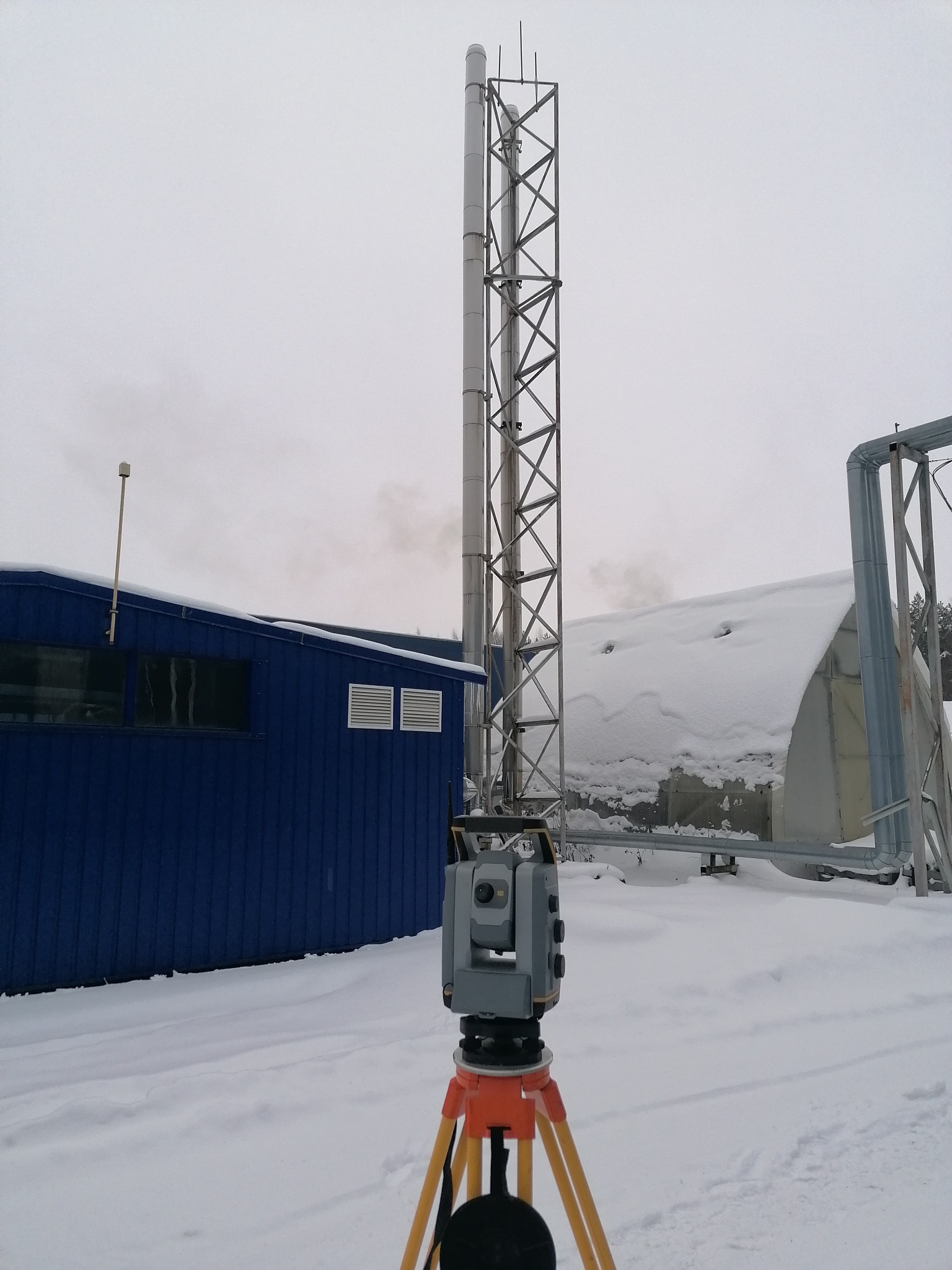 Szary teodolit na pomarańczowym statywie ustawiony na śniegu, w tle niebieski budynek i wysoki komin z metalową konstrukcją, pochmurne zimowe niebo.