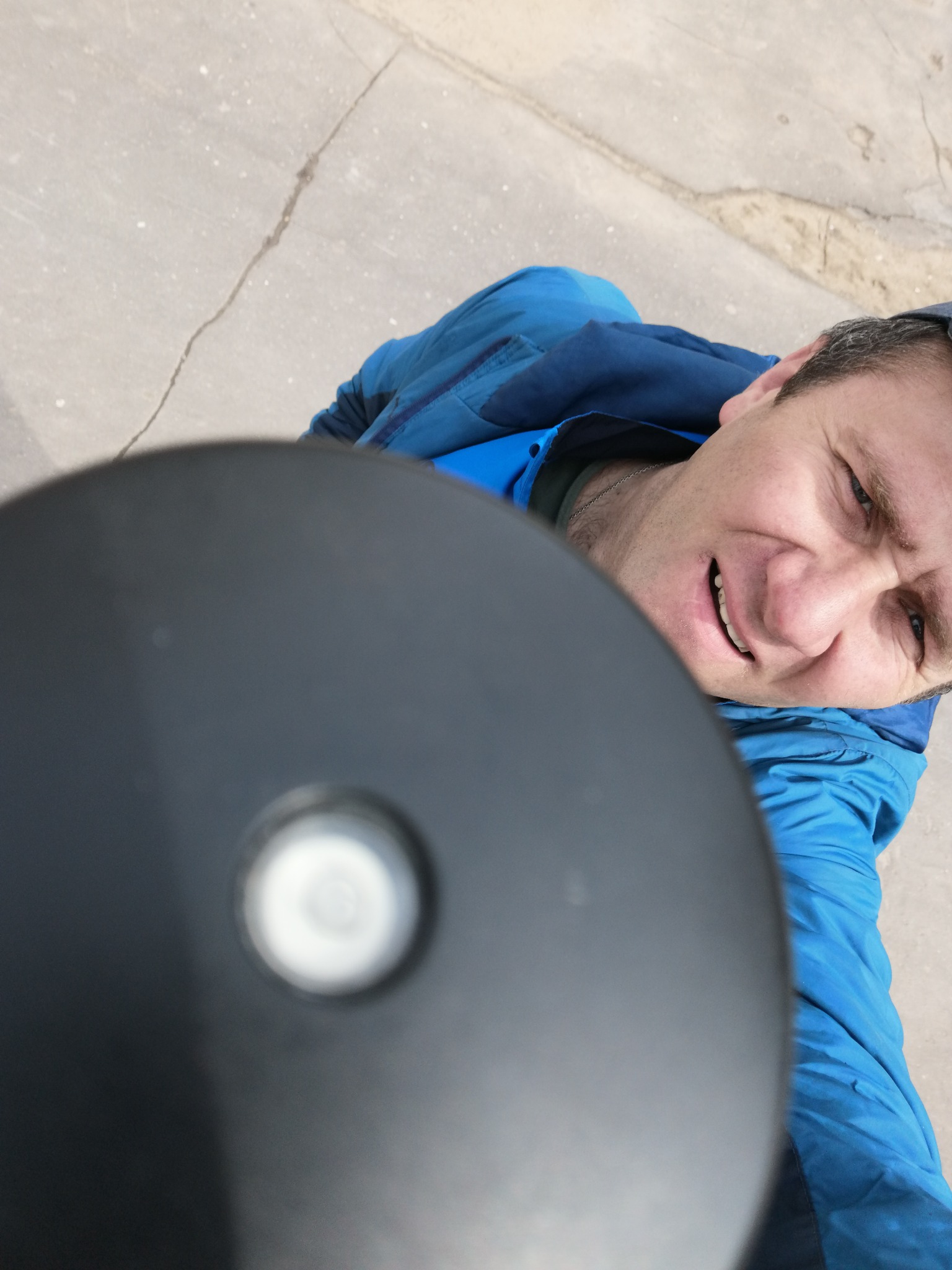 Mężczyzna w niebieskiej kurtce uśmiecha się, trzymając w dłoni niwelator optyczny podczas pracy na zewnątrz.