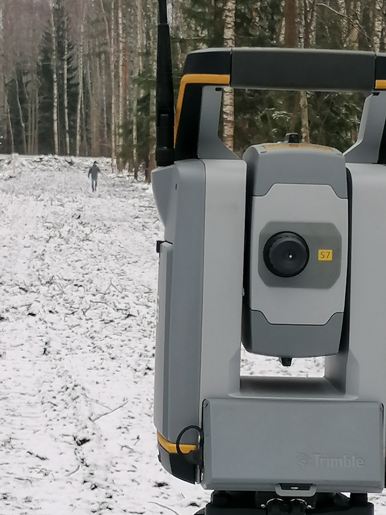 Szary teodolit Trimble S7 z anteną na tle zimowego krajobrazu z zalesionym terenem i oddalającą się postacią w tle.