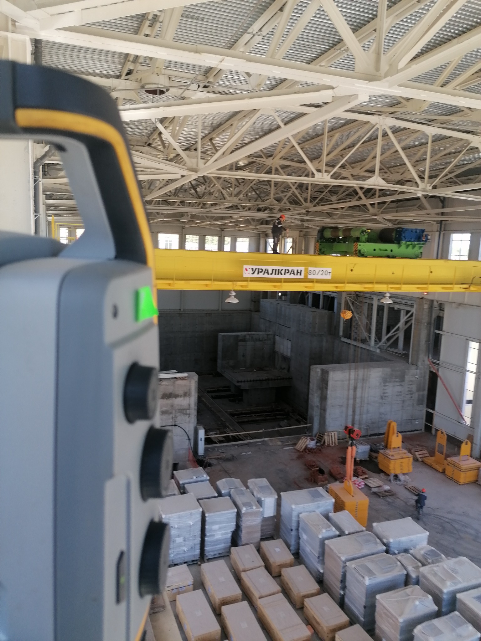 Nowoczesny teodolit z zieloną diodą w hali produkcyjnej, w tle suwnica z operatorem i przygotowane elementy konstrukcyjne.