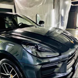 Błyszczący, ciemny Porsche Macan po umyciu, z otwartymi drzwiami i widocznymi kroplami wody na karoserii, w jasnym pomieszczeniu z folią ochronną na ścianach.