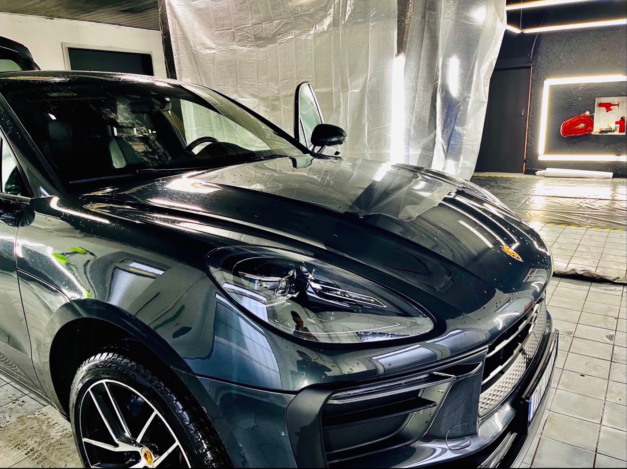 Błyszczący, ciemny Porsche Macan po umyciu, z otwartymi drzwiami i widocznymi kroplami wody na karoserii, w jasnym pomieszczeniu z folią ochronną na ścianach.