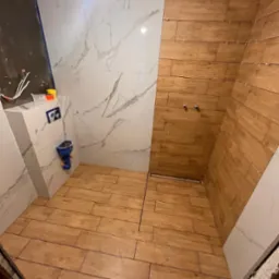 Wnętrze łazienki w trakcie remontu: położone drewnopodobne płytki na podłodze i ścianie prysznica, białe płytki z marmurowym wzorem na pozostałych ścianach, widoczne krzyżyki dystansowe między...