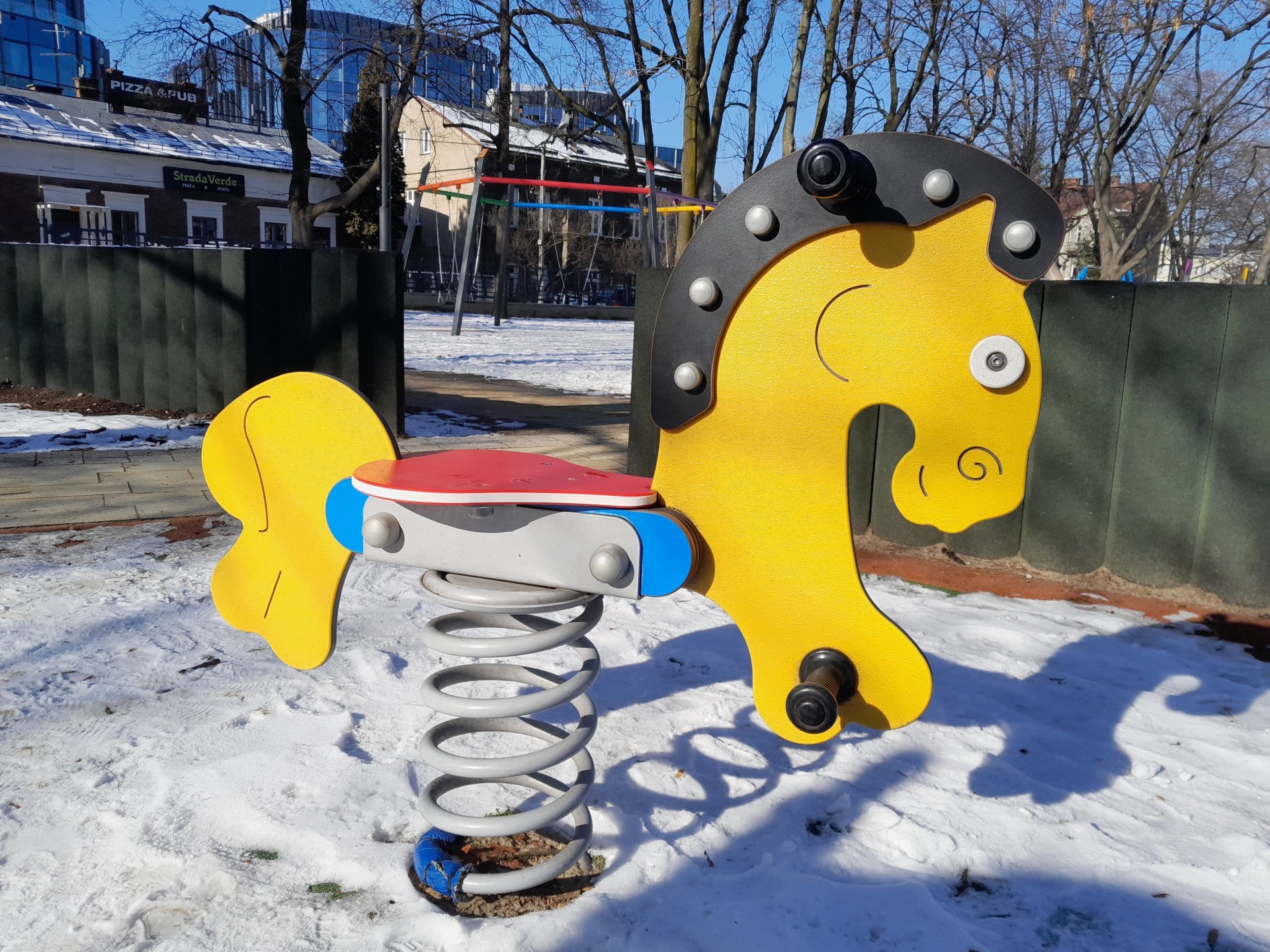 Żółty koń na sprężynie z czerwonym siedziskiem na zimowym placu zabaw, w tle budynki i drzewa bez liści.