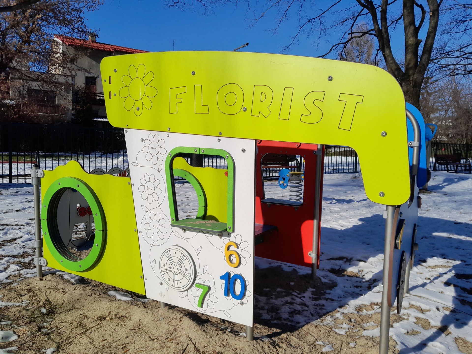 Kolorowa konstrukcja zabawowa z napisem 'FLORIST' i elementami edukacyjnymi, takimi jak liczby i rysunki kwiatów, umieszczona na piaszczystym podłożu z resztkami śniegu w tle.