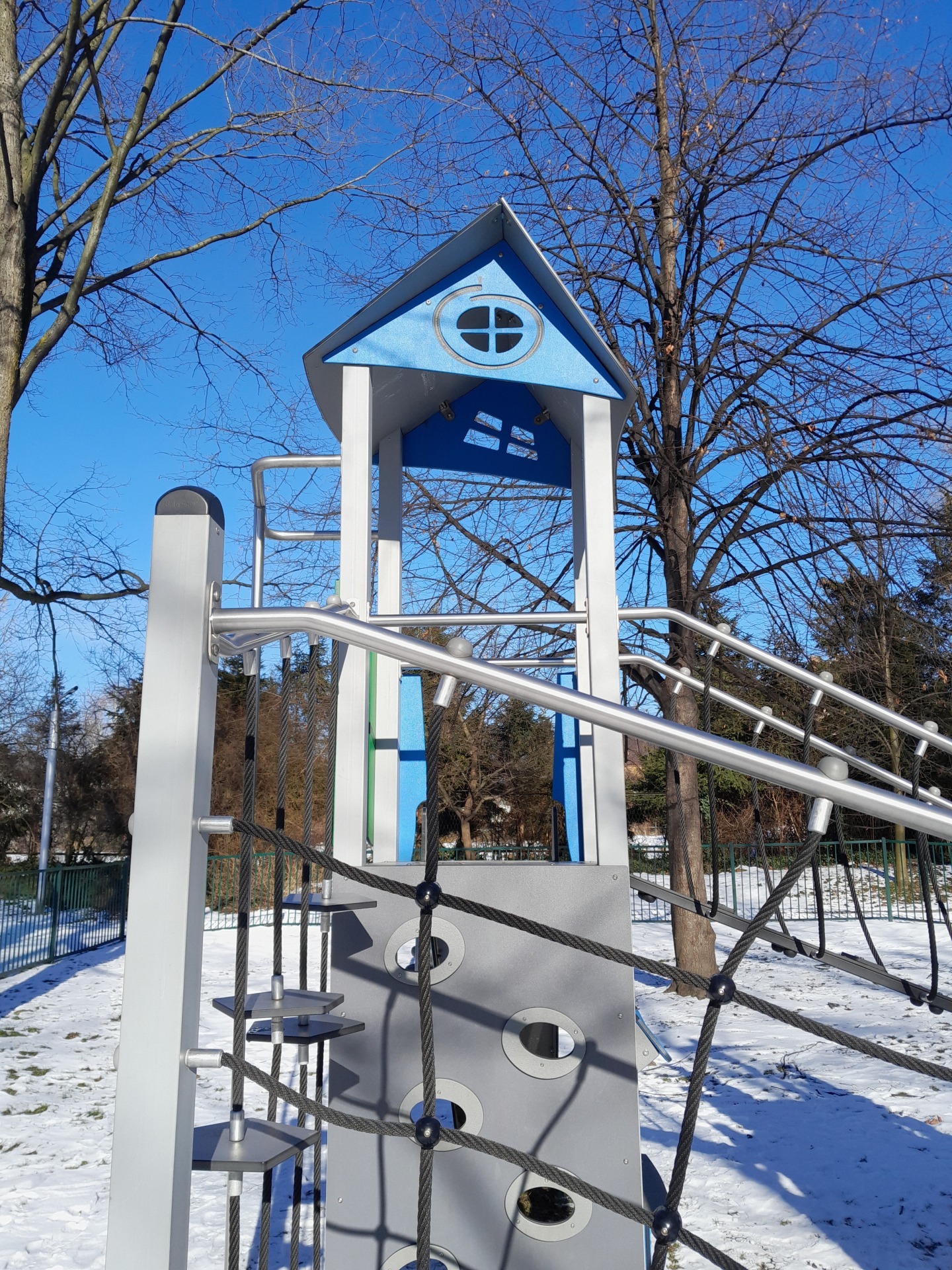 Wieża ze schodami linowymi i zjeżdżalnią na placu zabaw, zimowy krajobraz z drzewami bez liści i śniegiem.