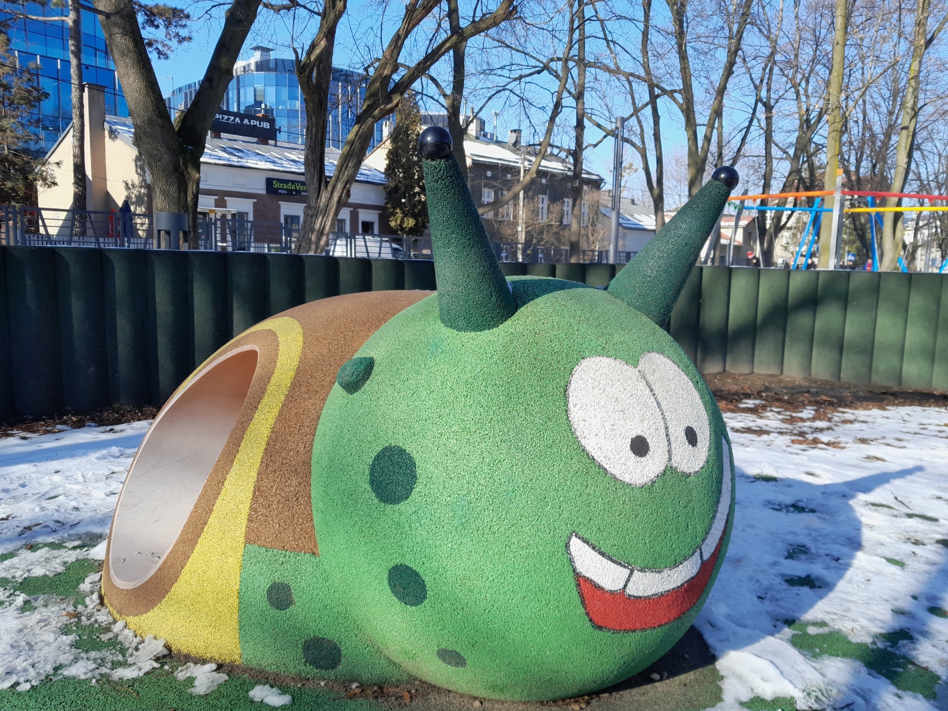 Duży, zielony ślimak z tworzywa sztucznego z uśmiechniętą buzią i czułkami na placu zabaw, z tunelem w skorupie, otoczony śniegiem i budynkami w tle.