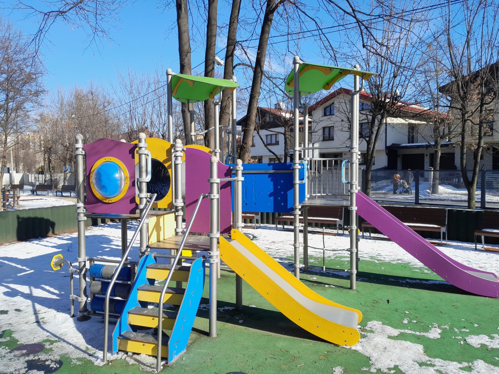 Kolorowy zestaw zabawowy z dwiema zjeżdżalniami (żółtą i fioletową), schodkami i elementami do wspinaczki, na zielonej nawierzchni z resztkami śniegu, w tle budynki mieszkalne i drzewa bez liści.