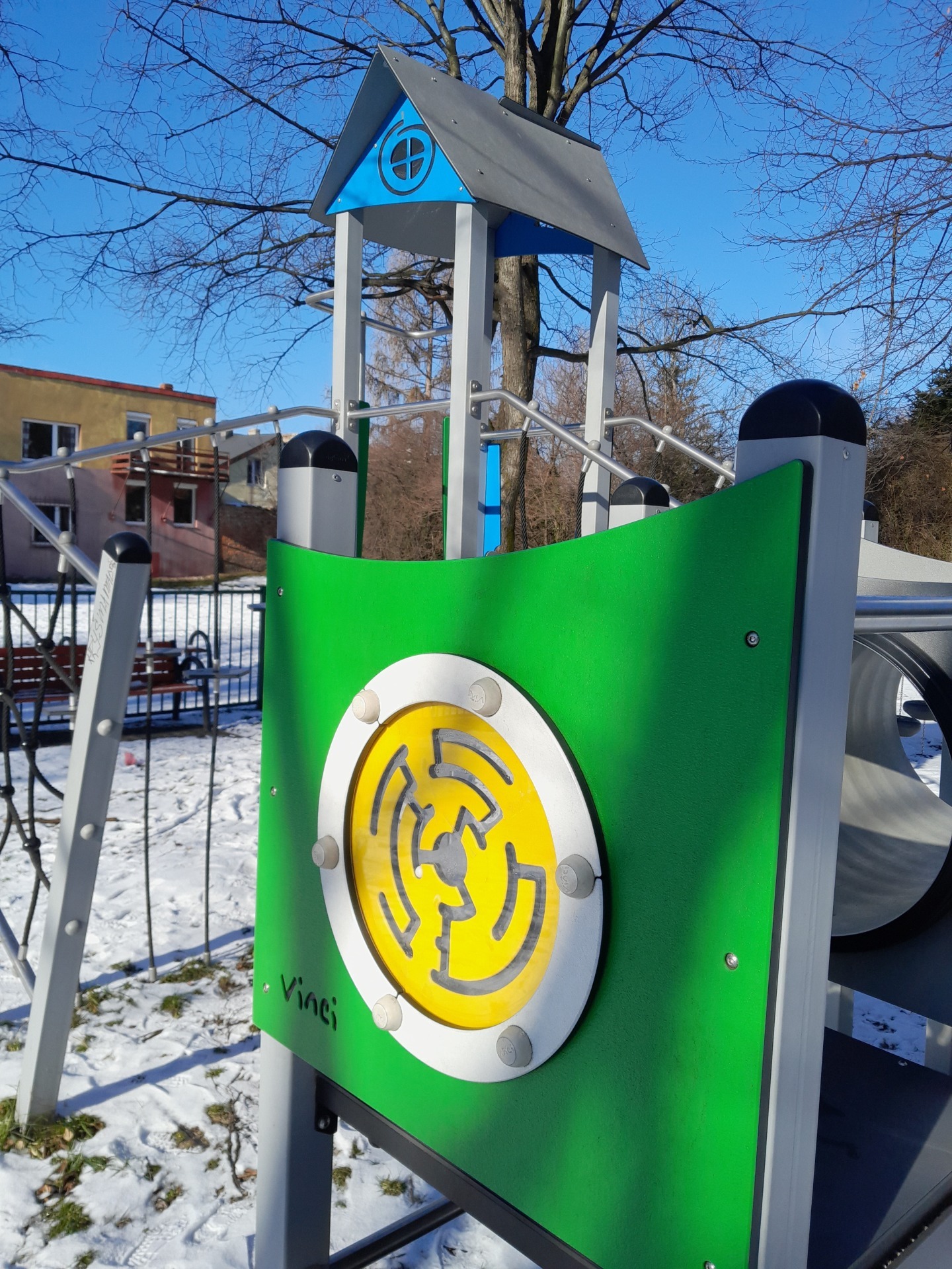 Fragment konstrukcji placu zabaw z zielonym panelem z żółtym labiryntem i napisem Vinci, na tle zimowego krajobrazu.