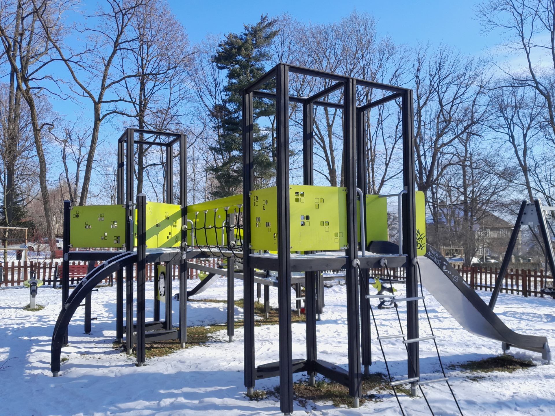 Wieloelementowa konstrukcja zabawowa z metalu i zielonych paneli na zasnieżonym placu, widok na drabinkę, zjeżdżalnię i most linowy.