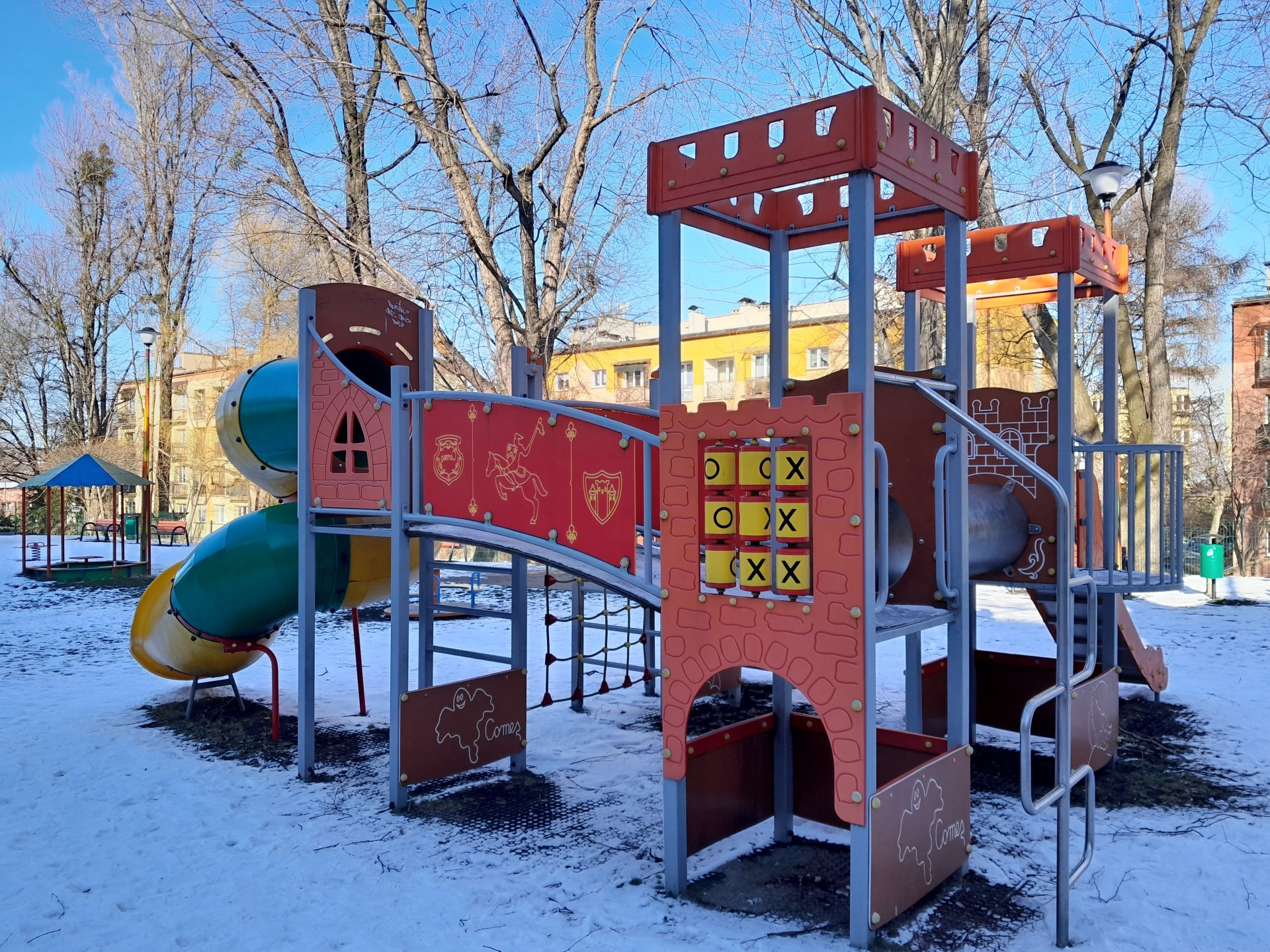 Kolorowy zestaw zabawowy na zewnątrz z tunelem, zjeżdżalnią i elementami do wspinaczki, otoczony śniegiem w słoneczny dzień.