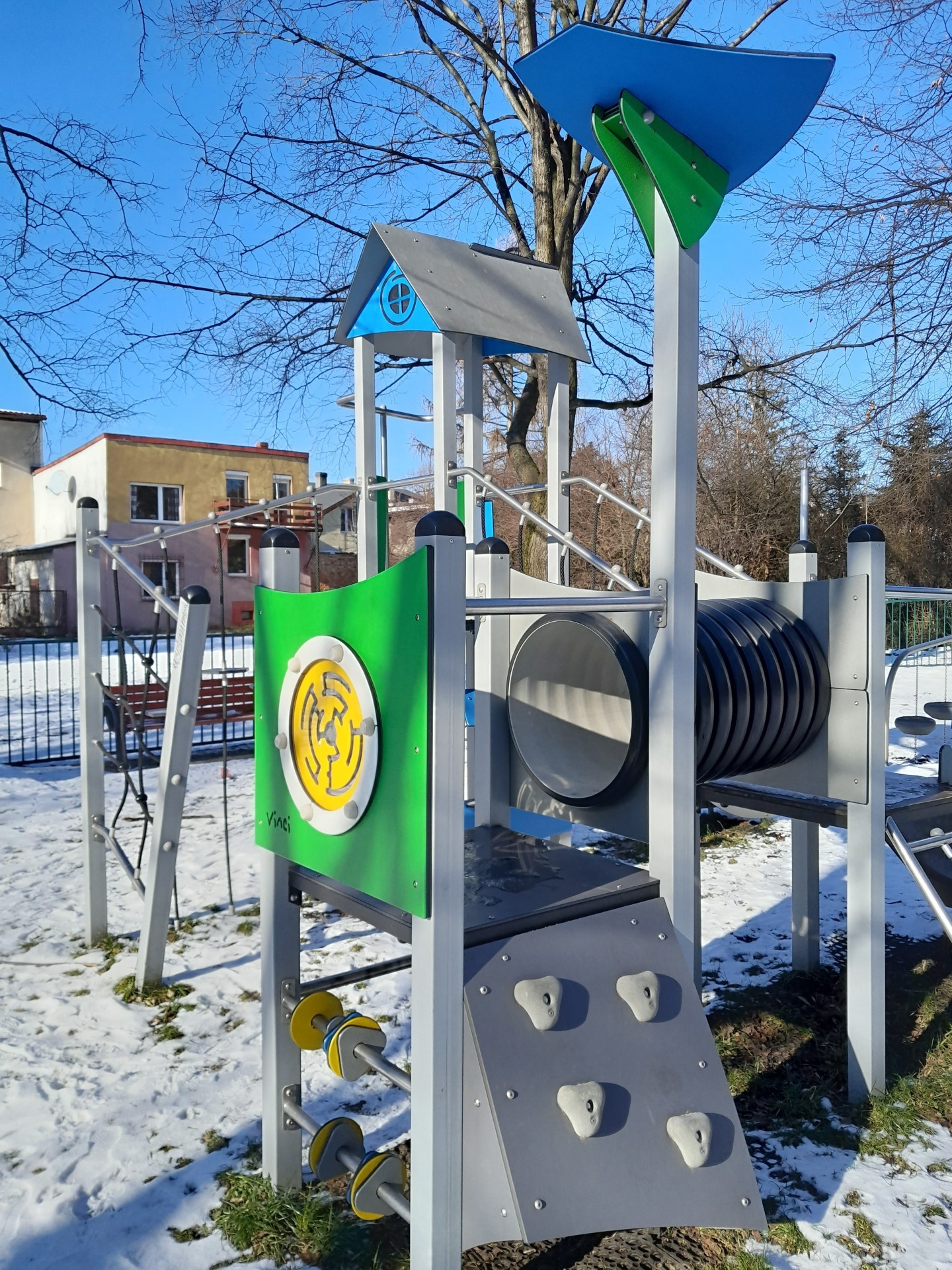 Wieloelementowa konstrukcja zabawowa z tunelem, ścianką wspinaczkową i panelem edukacyjnym z labiryntem, na zimowym tle z fragmentem osiedla