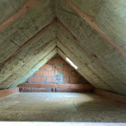 Poddasze z ociepleniem z wełny mineralnej, widoczne krokwie, sznurki mocujące izolację i fragment ściany z cegły dziurawki.