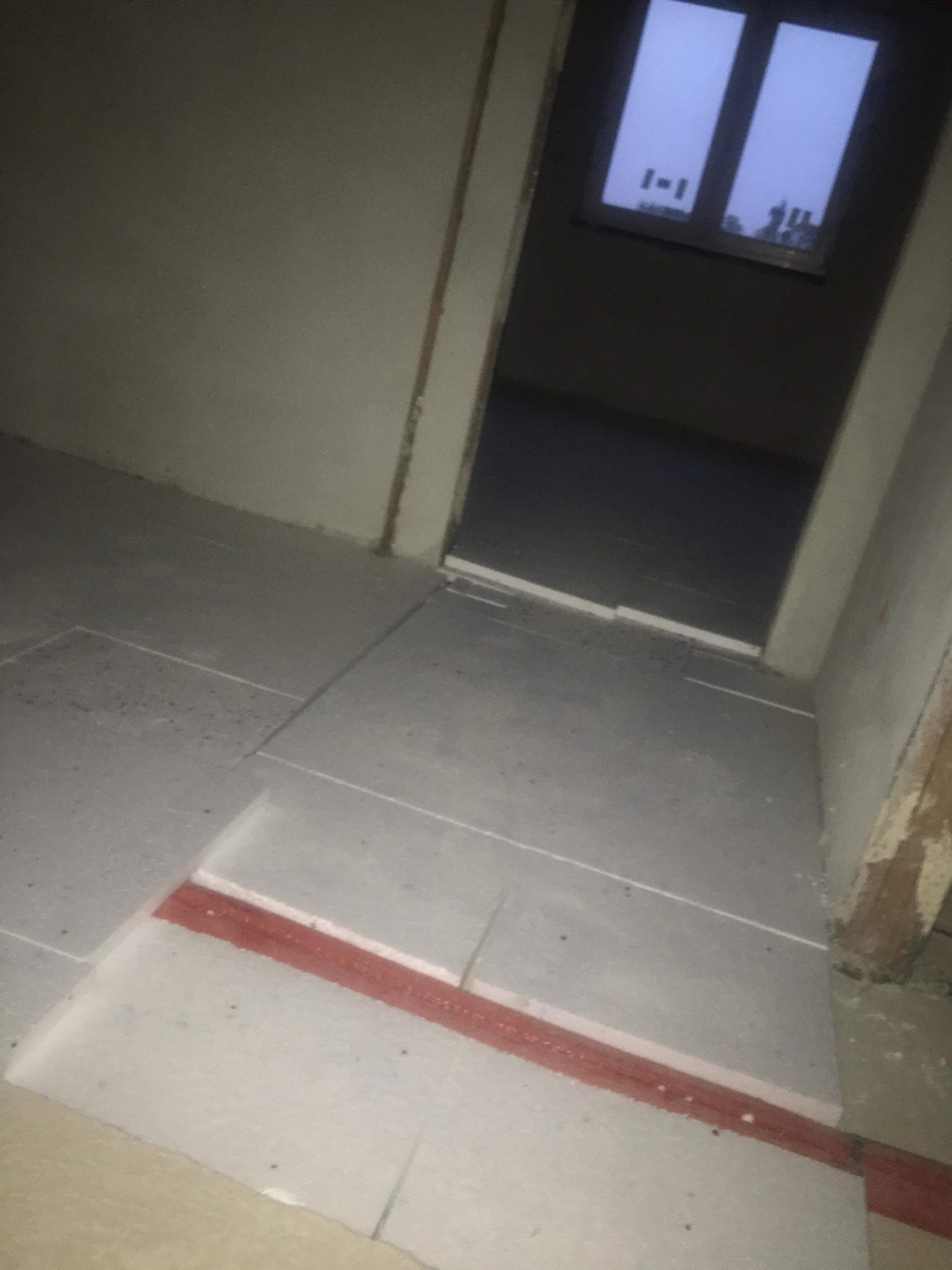 Przygotowanie podłoża pod posadzkę: płyty styropianowe z czerwoną taśmą łączącą, widok na surowe ściany i okno w tle.