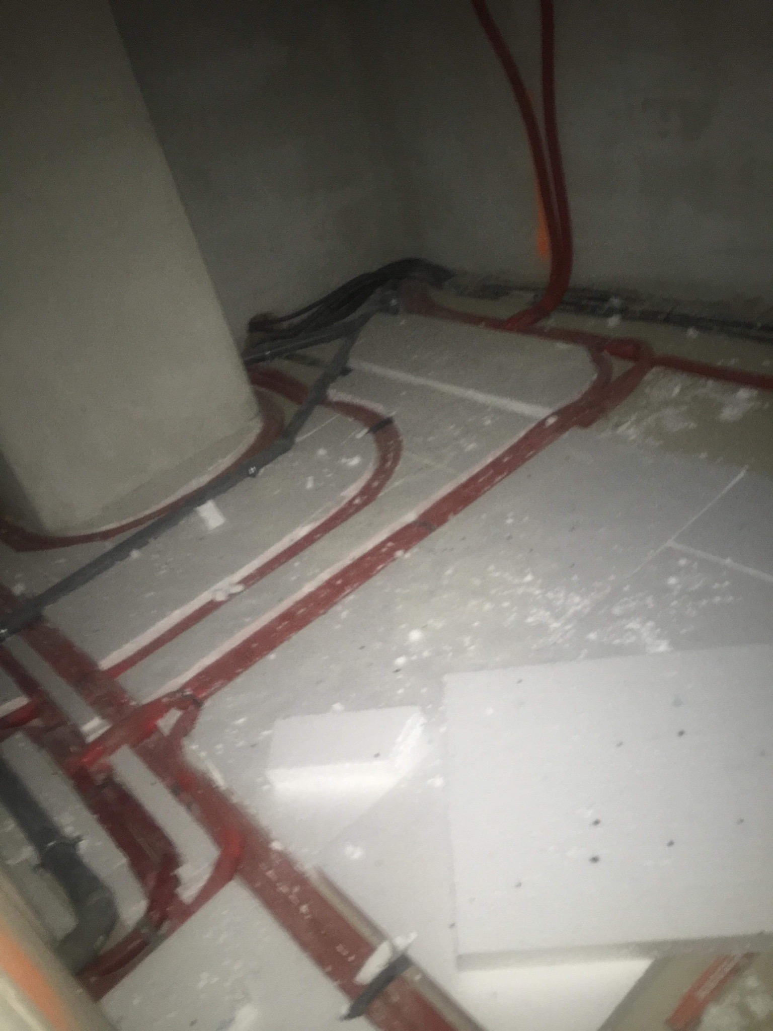 Instalacja ogrzewania podłogowego z czerwonych rur na białym styropianie, widoczne czarne kable elektryczne w rogu pomieszczenia, surowy beton na ścianach.