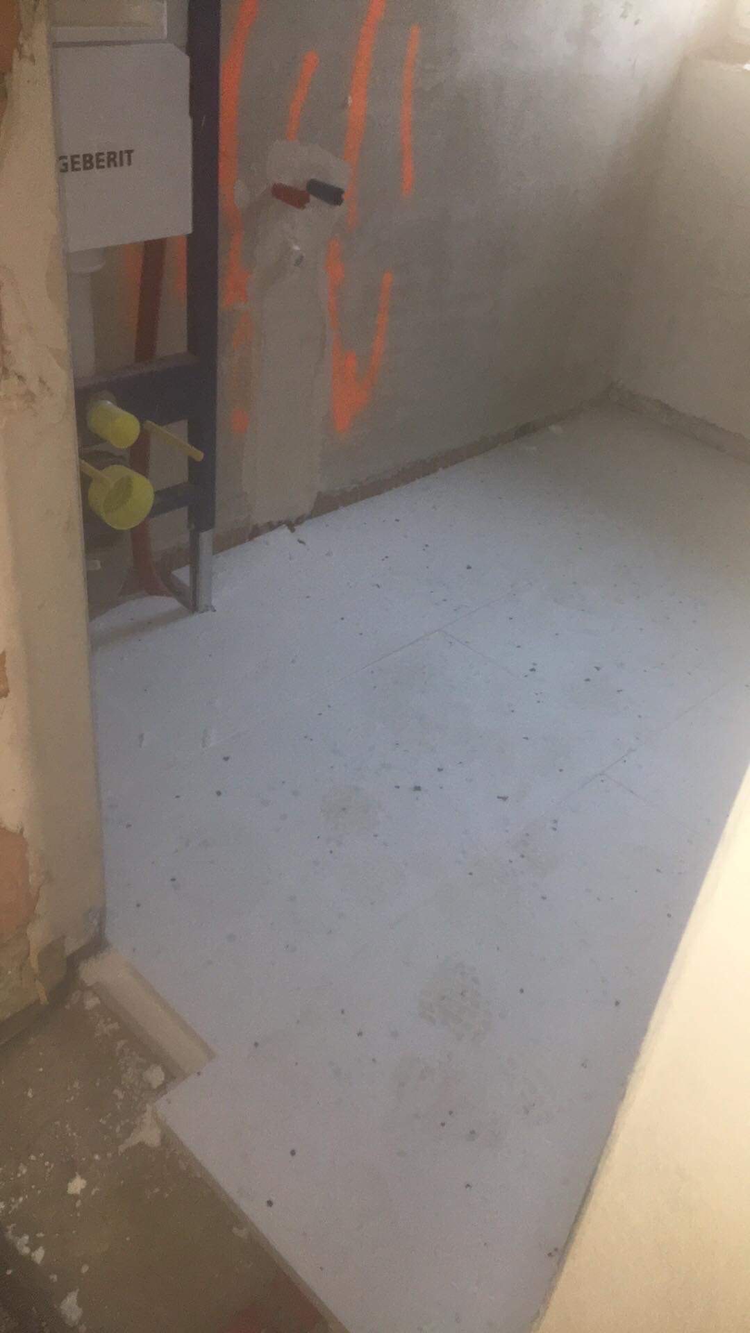 Surowy stan łazienki w trakcie prac: widoczne instalacje Geberit, ściany z pomarańczowymi oznaczeniami, podłoga z wylewką samopoziomującą przed położeniem płytek.