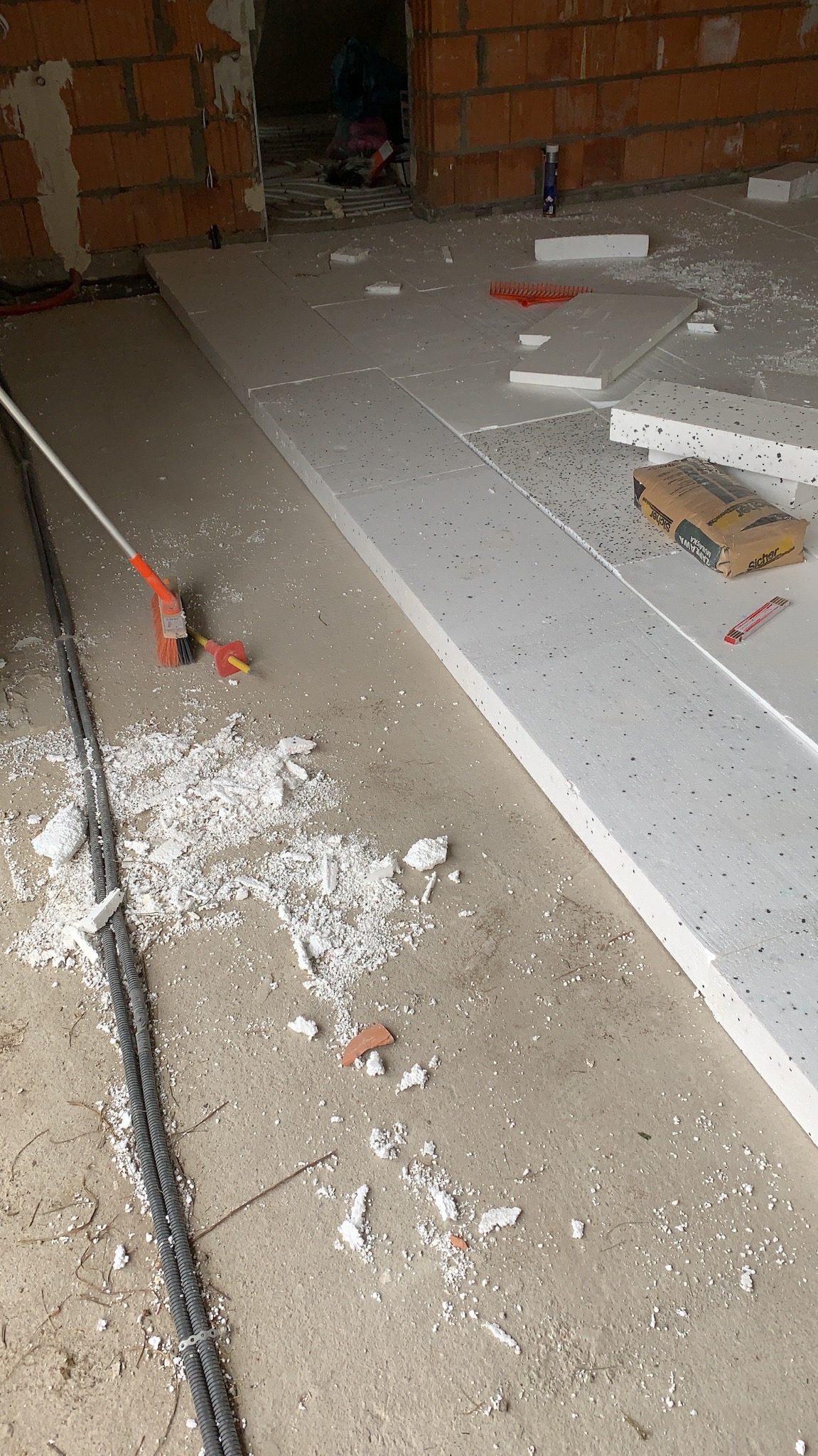 Układanie białych płyt styropianowych na podłodze, widoczne przewody w peszlu, resztki styropianu, paczka kleju i nóż do cięcia.