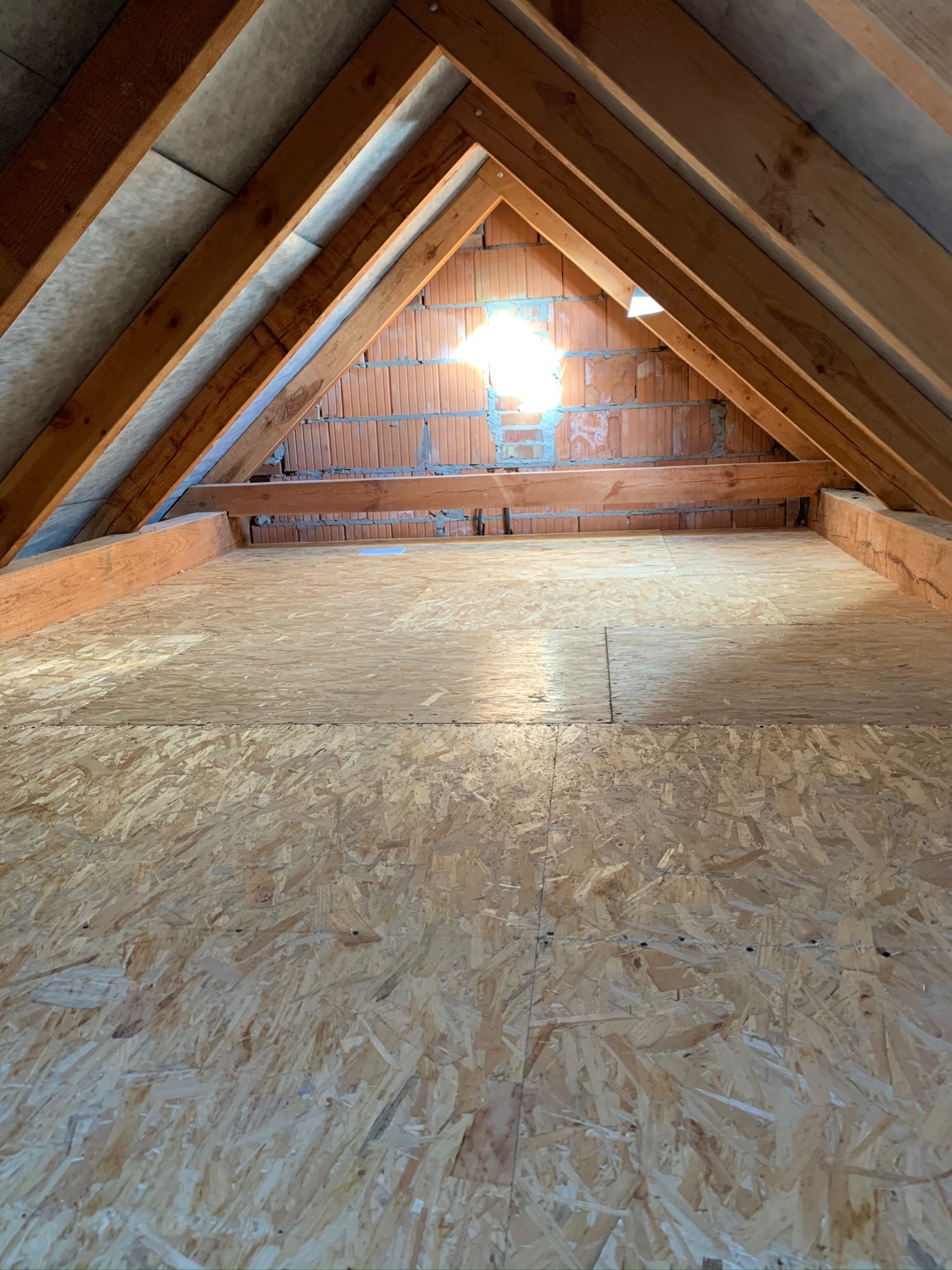 Poddasze z drewnianą konstrukcją więźby dachowej, oknem w szczycie i podłogą wyłożoną płytami OSB.