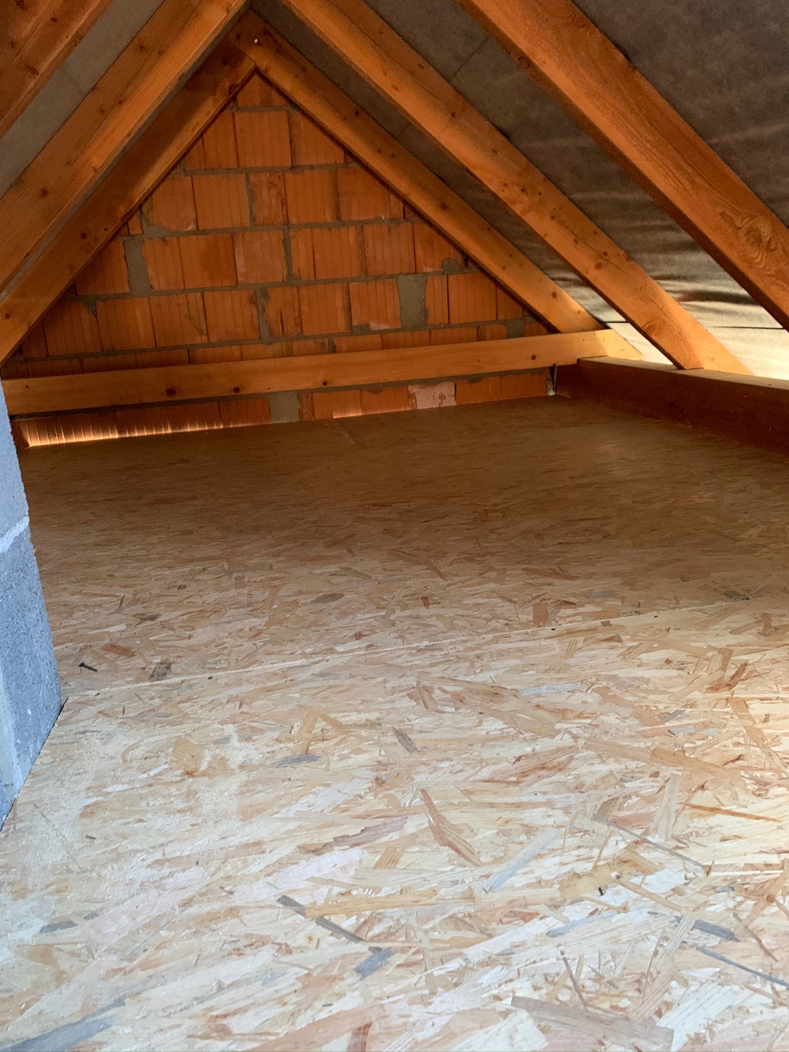 Poddasze z widoczną konstrukcją dachu z drewnianych belek, ceglaną ścianą szczytową i podłogą z płyt OSB.