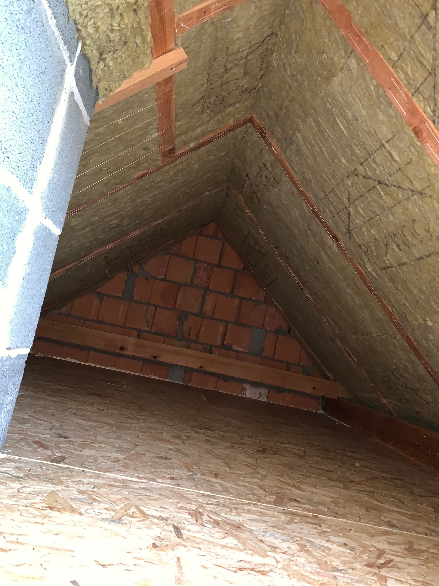 Poddasze w trakcie ocieplania, widoczna wełna mineralna między krokwiami, murowana ściana szczytowa z cegły oraz płyta OSB na podłodze.