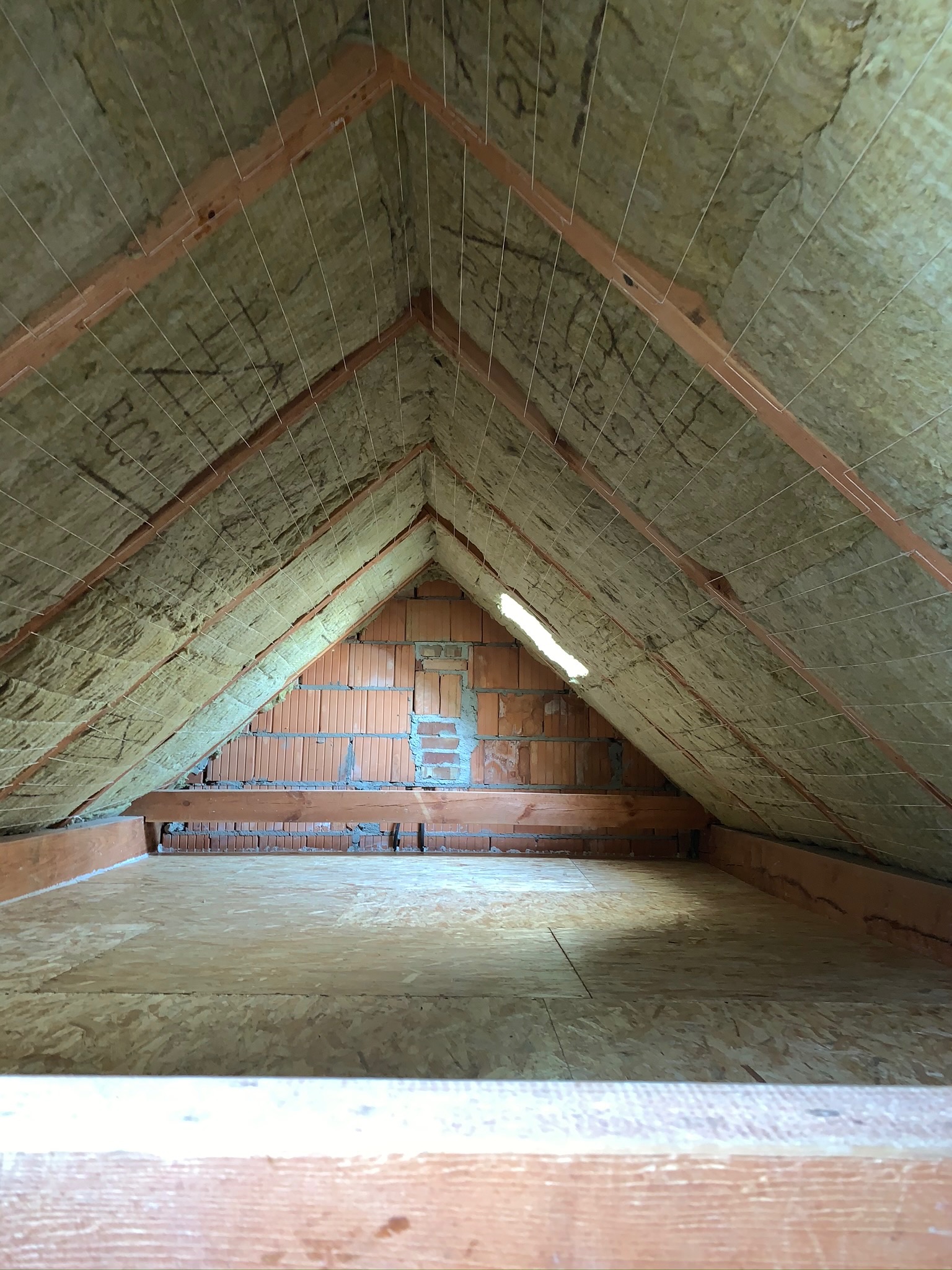 Poddasze z ociepleniem z wełny mineralnej, widoczne krokwie, sznurki mocujące izolację i fragment ściany z cegły dziurawki.