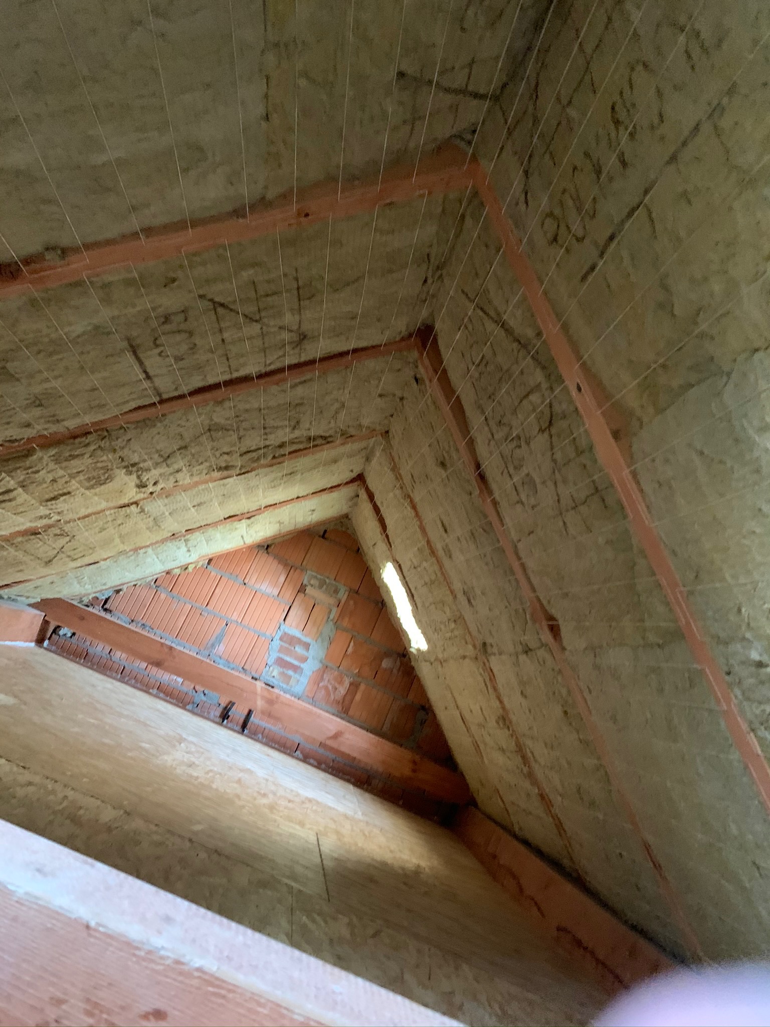 Skośny widok na ocieplone wełną mineralną poddasze z widoczną konstrukcją drewnianą, ceglanym kominem i siatką podtrzymującą izolację.