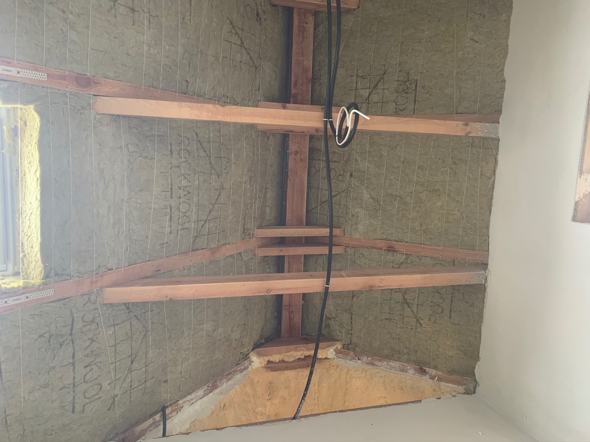 Poddasze z ociepleniem z wełny mineralnej, drewnianą konstrukcją nośną i napiętymi drutami mocującymi, widoczne przewody elektryczne i fragment okna.