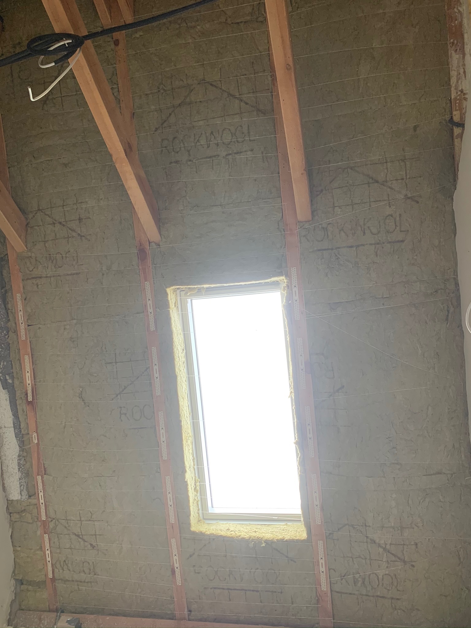 Poddasze z oknem dachowym w trakcie izolacji wełną mineralną Rockwool, widoczne drewniane krokwie, listwy mocujące i naciągnięte druty podtrzymujące izolację.