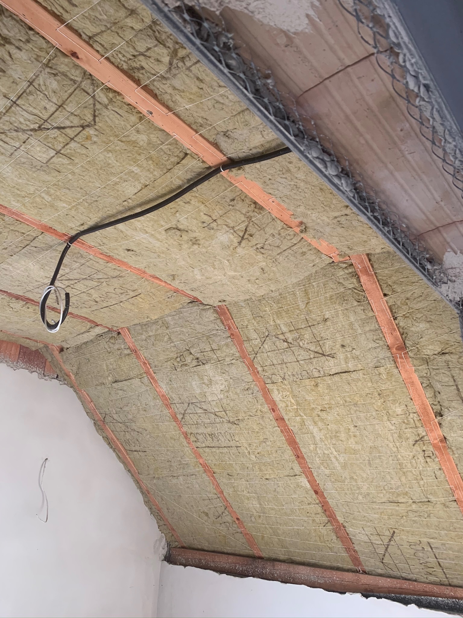 Izolacja poddasza wełną mineralną, widoczne drewniane belki konstrukcyjne, siatka zabezpieczająca i przewody elektryczne w trakcie montażu.