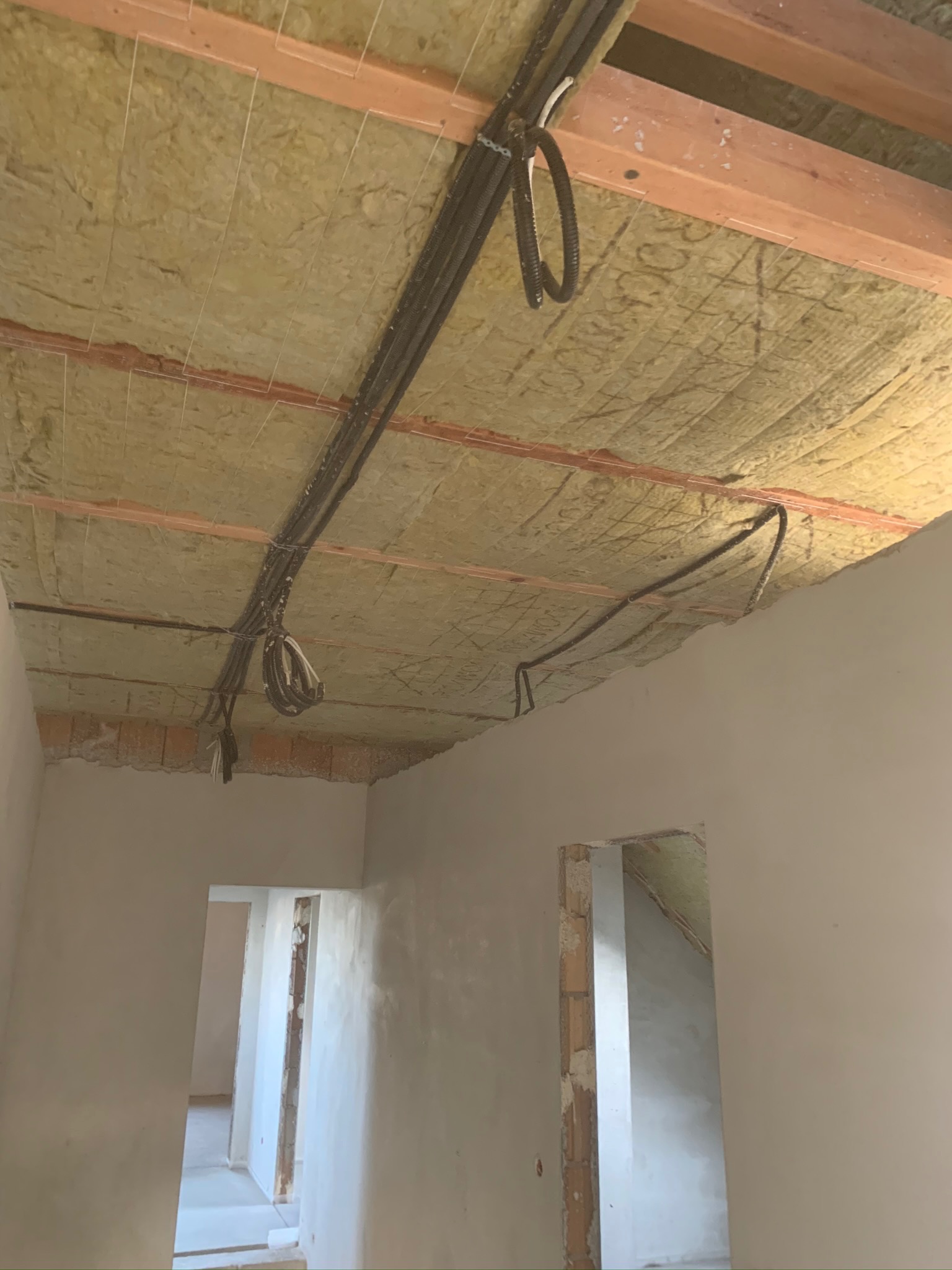 Poddasze ocieplone wełną mineralną z widocznymi kablami elektrycznymi i drewnianą konstrukcją nośną, ściany w trakcie szpachlowania, w tle widoczne przejścia do innych pomieszczeń.