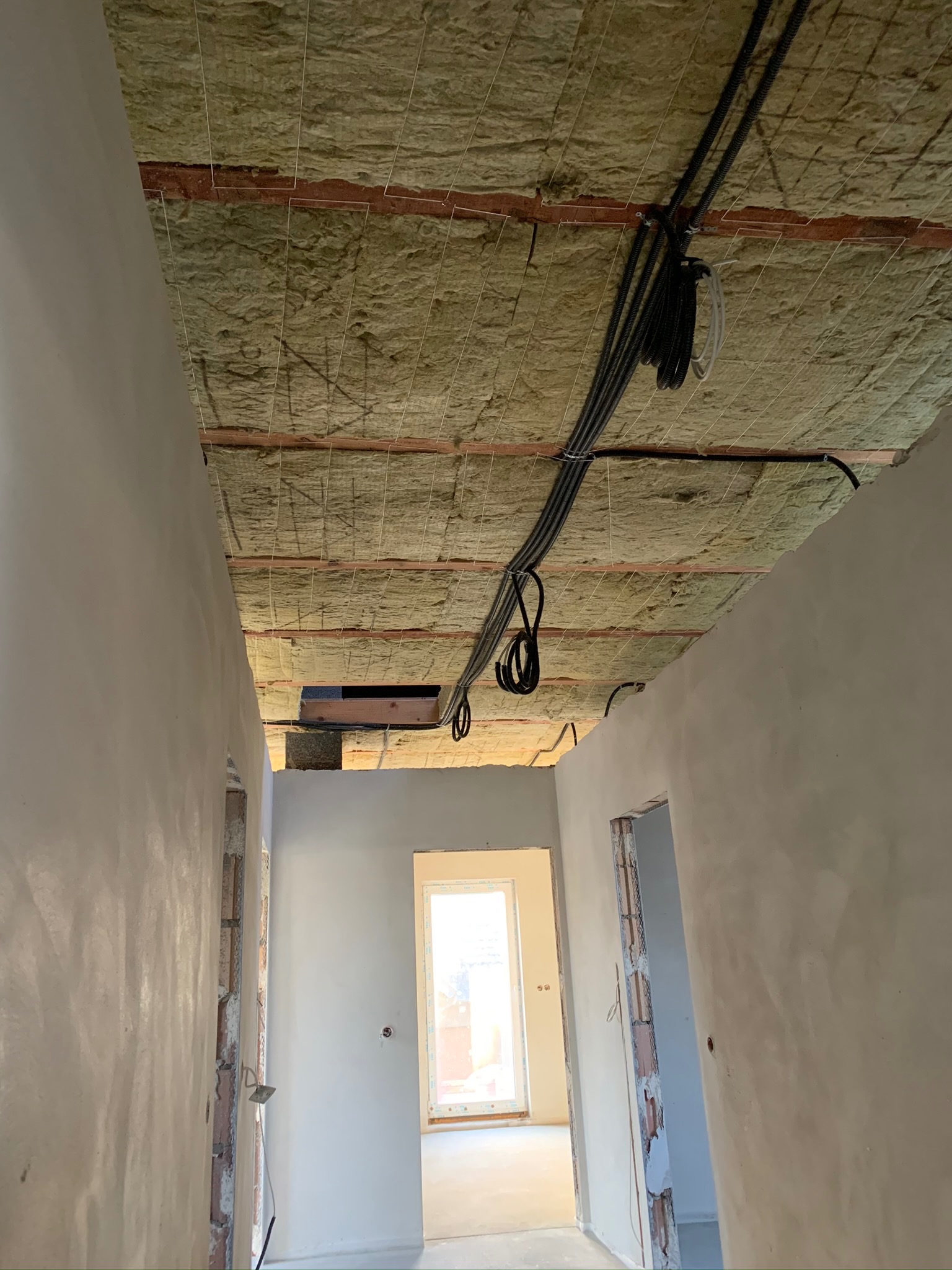 Poddasze w trakcie ocieplania wełną mineralną, widoczne przewody elektryczne mocowane do drewnianej konstrukcji stropu, ściany w stanie surowym po nałożeniu tynku.