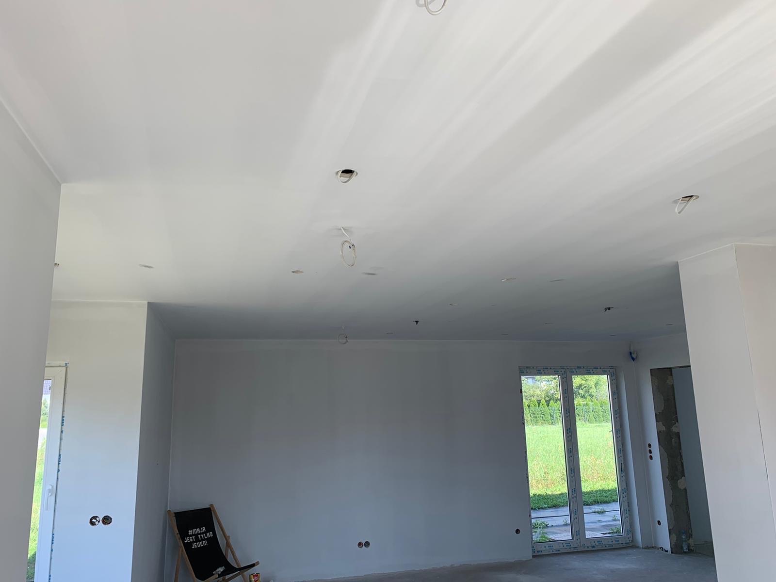 Wnętrze świeżo pomalowanego pokoju z widocznymi otworami na oświetlenie w suficie, z krzesłem plażowym z napisem opartym o ścianę oraz oknem balkonowym z widokiem na zieleń.