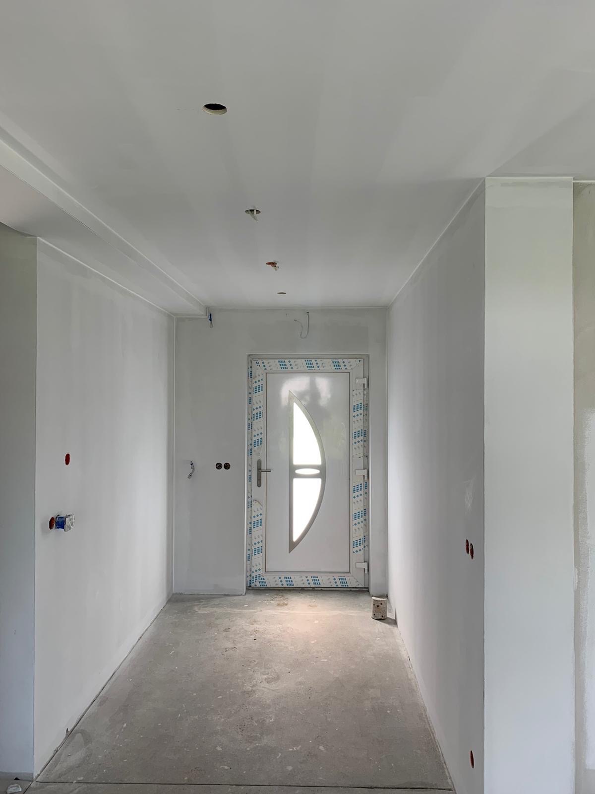 Korytarz w trakcie prac wykończeniowych, świeżo pomalowane ściany na biało, widoczne puszki elektryczne bez osprzętu, białe drzwi z owalnym przeszkleniem zabezpieczone folią ochronną na ościeżnicy,...