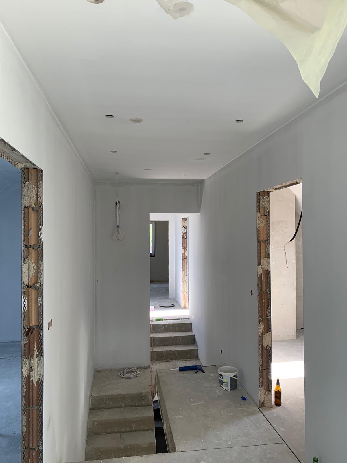 Korytarz w trakcie prac wykończeniowych: ściany pomalowane na szaro, widoczne niedokończone ościeżnice, schody betonowe, puszka farby i butelka po piwie na podłodze, folia malarska przy suficie.