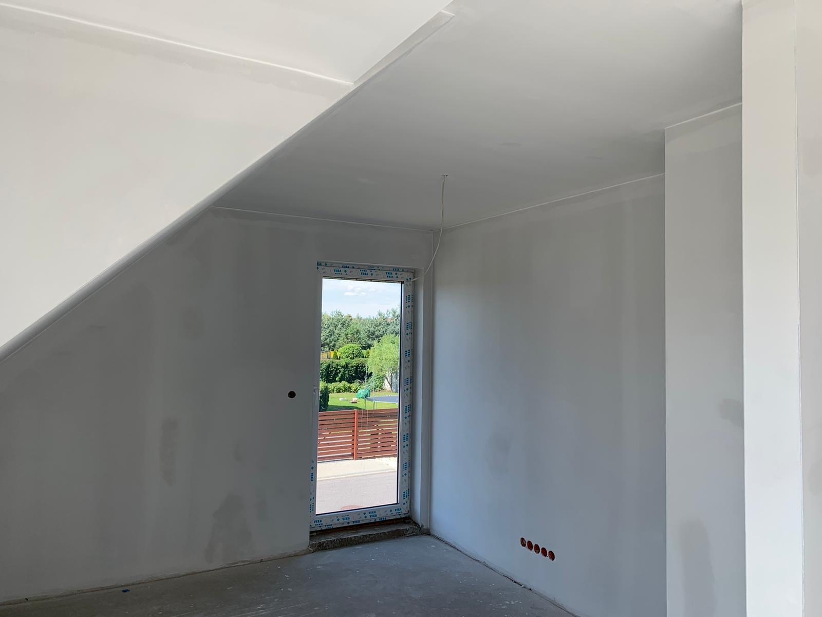 Wnętrze pokoju na poddaszu w trakcie prac wykończeniowych: szpachlowane ściany, okno z widokiem na zewnątrz, widoczne punkty elektryczne, surowa podłoga, skos dachu.