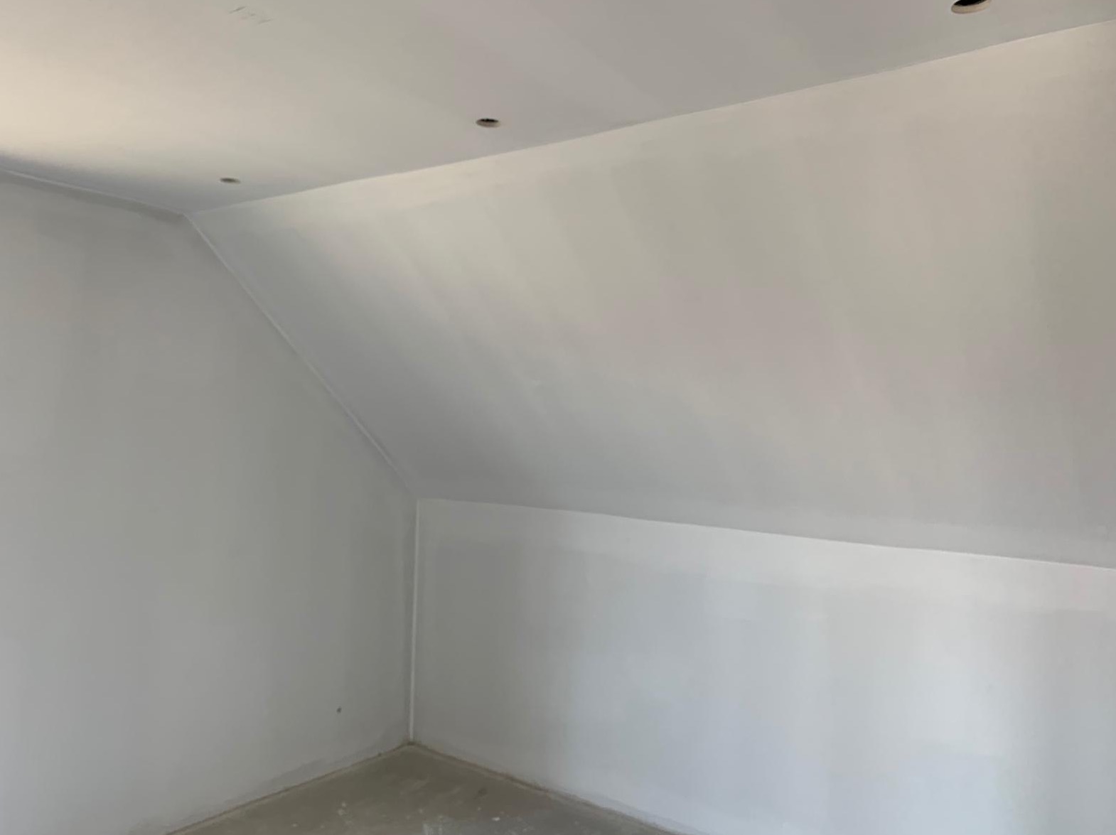 Pomalowane na biało poddasze ze skosami, widoczne otwory na oświetlenie w suficie, szary beton na podłodze.