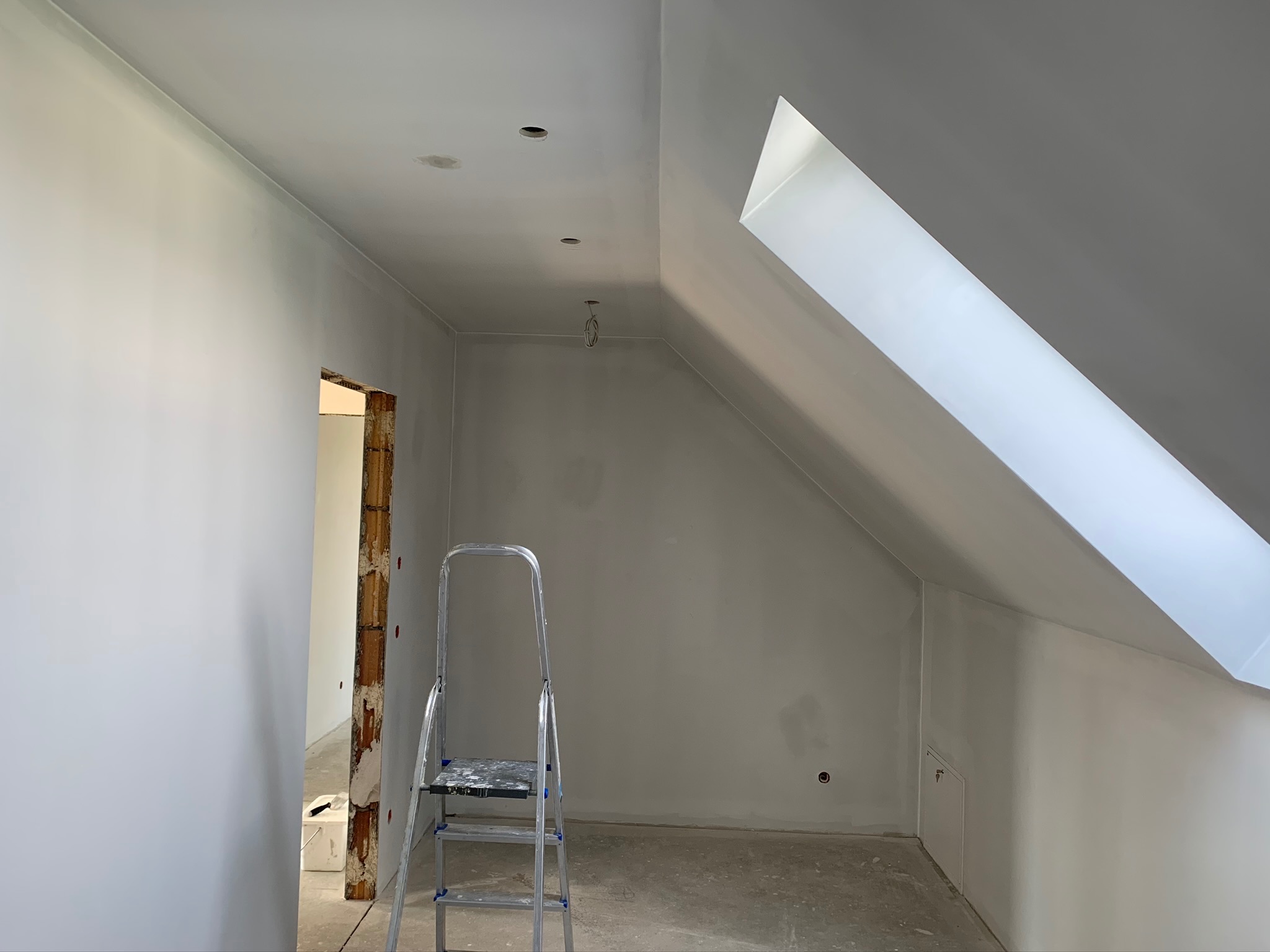 Poddasze po malowaniu na biało, widoczna drabina aluminiowa, okno dachowe i surowa futryna drzwiowa.