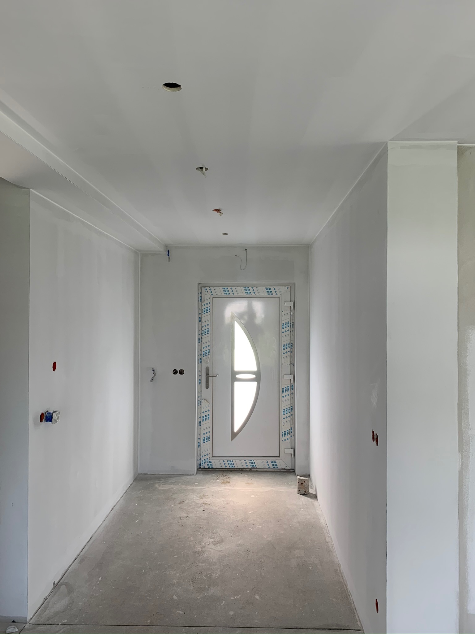 Korytarz w trakcie prac wykończeniowych: świeżo pomalowane ściany na biało, surowy beton na podłodze, otwory na oświetlenie w suficie, białe drzwi z półokrągłym przeszkleniem zabezpieczone folią.