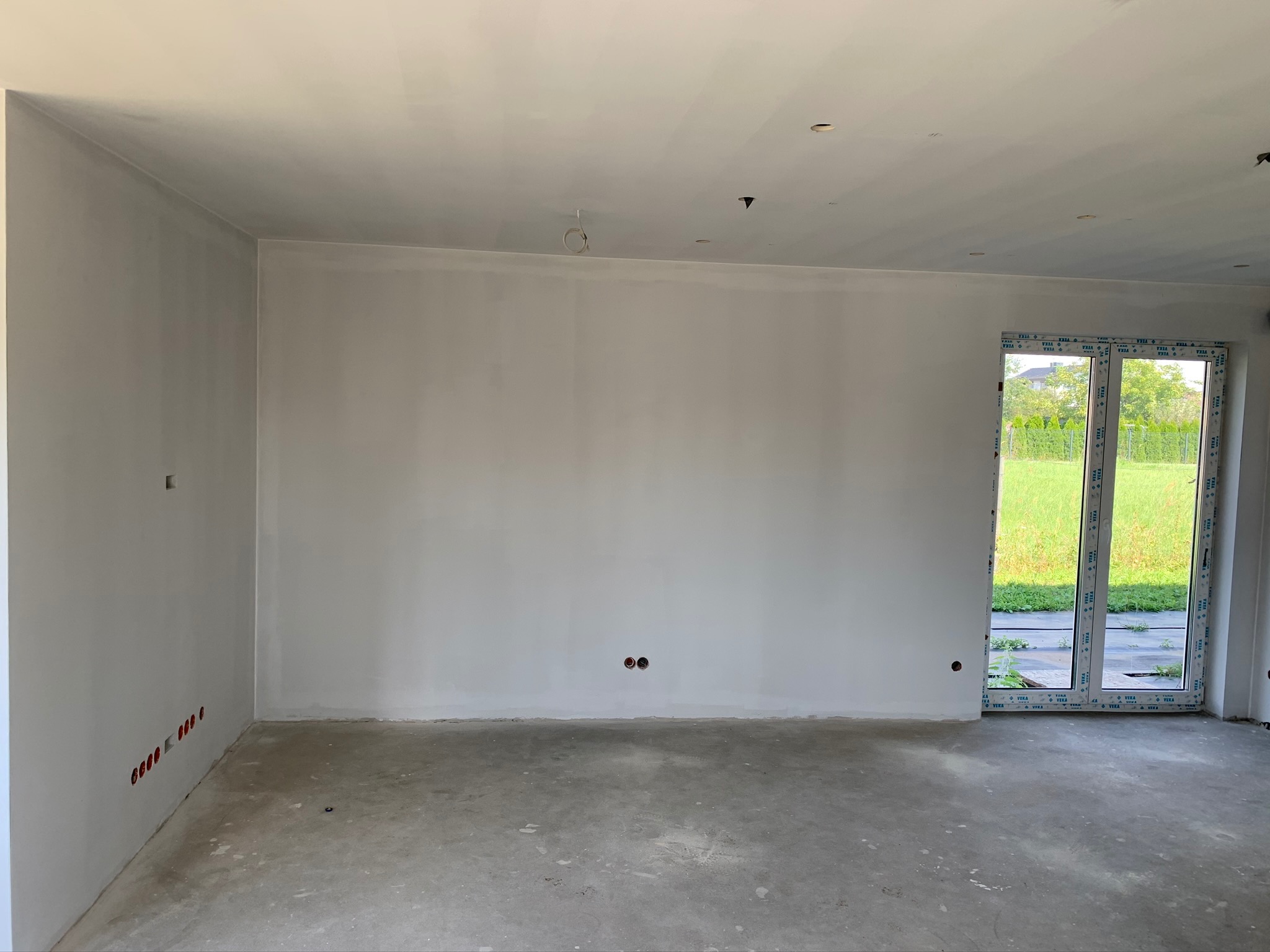 Surowe wnętrze z gładzią na ścianach, widoczne puszki elektryczne i okno balkonowe z widokiem na zieleń.