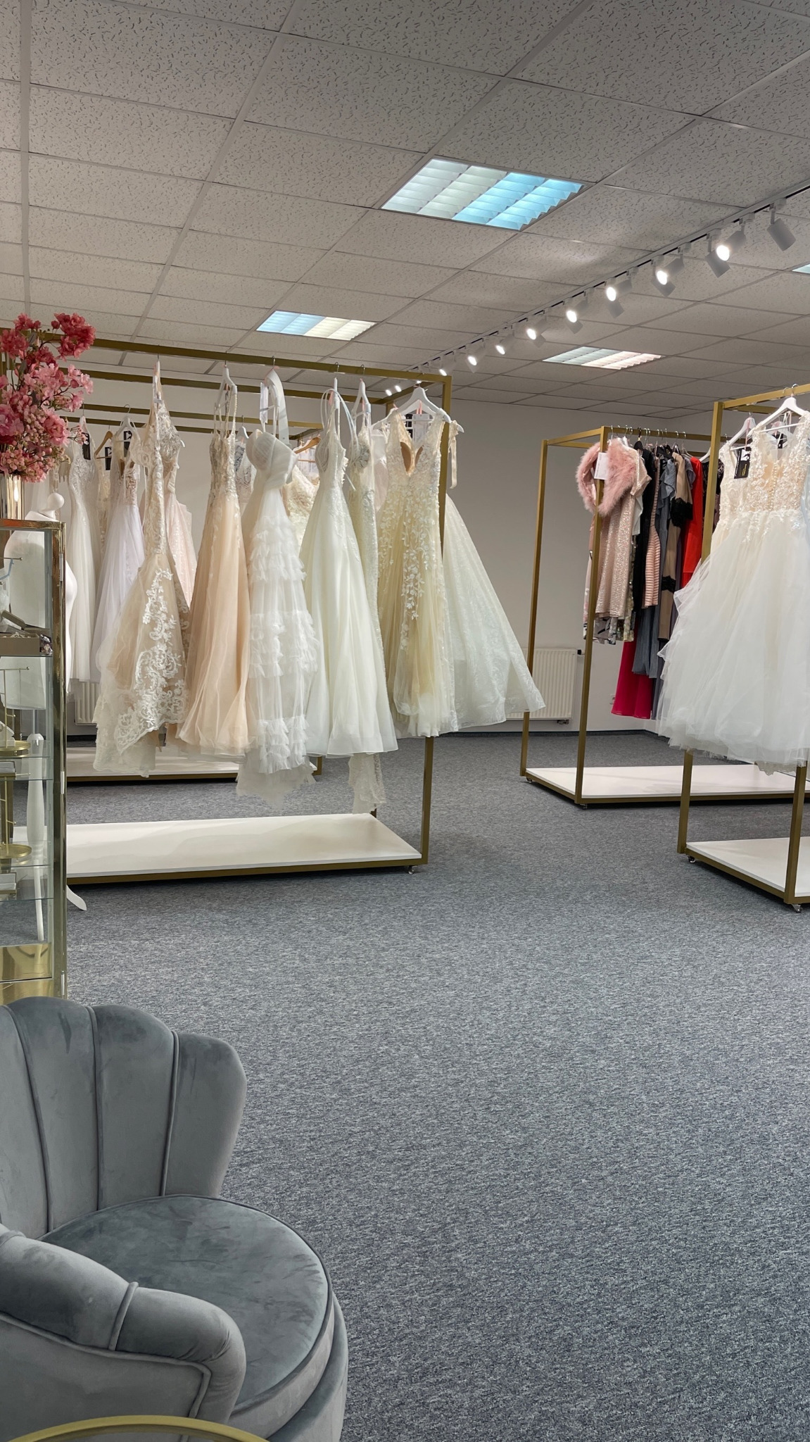 Salon sukien ślubnych z ekspozycją na złotych wieszakach, w tle różowe kwiaty i szary fotel.