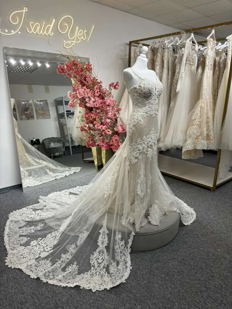 Koronkowa suknia ślubna typu rybka na manekinie, z długim trenem, w salonie z neonem 'I said Yes!' i dekoracją z różowych kwiatów.