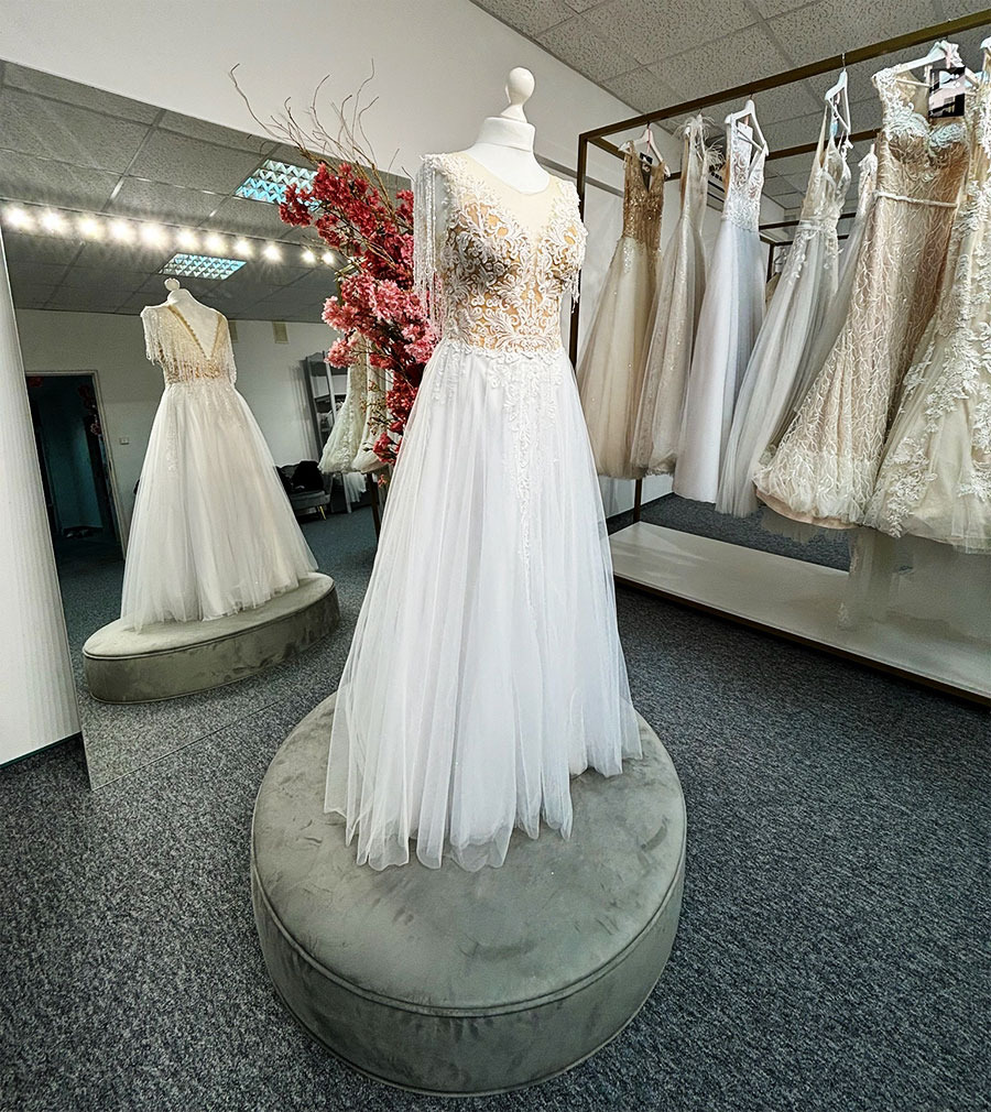 Salon sukien ślubnych: Manekin prezentuje białą suknię ślubną z koronkową górą na tle innych sukien i lustra.