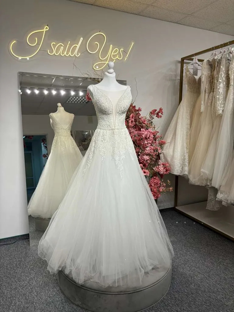 Elegancka suknia ślubna z koronkową górą i tiulowym dołem na manekinie w salonie, z neonowym napisem 'I said Yes!' w tle i bukietem różowych kwiatów obok.