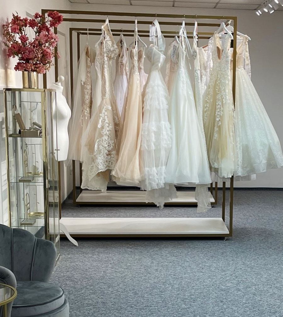 Salon sukien ślubnych: kolekcja delikatnych sukien w odcieniach bieli i ecru, wiszących na złotym wieszaku ekspozycyjnym, obok szklanej witryny z biżuterią i bukietem różowych kwiatów.