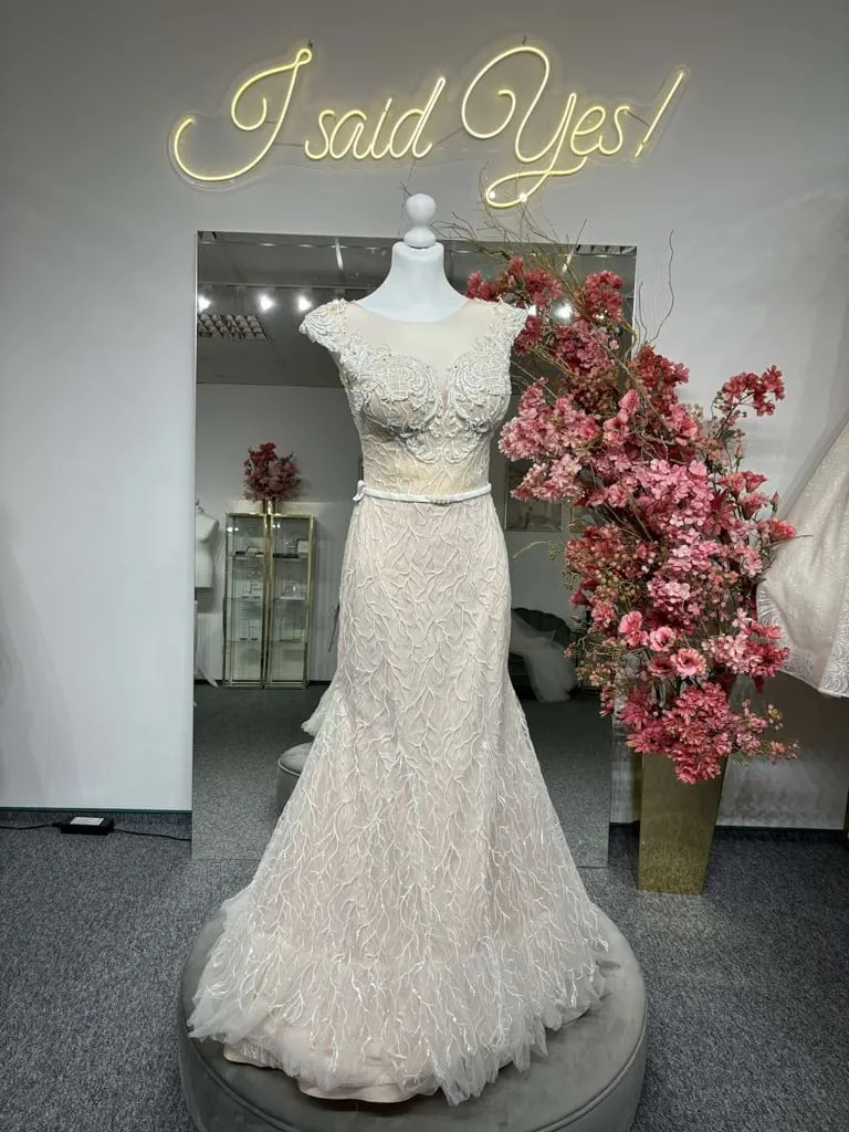 Elegancka suknia ślubna typu rybka z delikatnym koronkowym wzorem, prezentowana na manekinie w salonie, z neonowym napisem 'I said Yes!' w tle i dekoracją z różowych kwiatów.