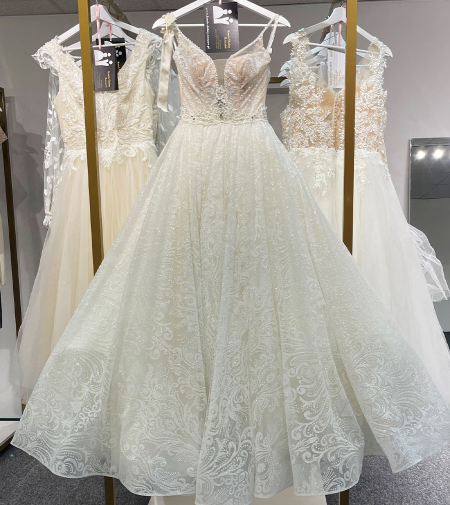 Biała suknia ślubna z koronkowym wzorem i zdobieniami na gorsecie, wisząca na wieszaku w salonie, obok inne suknie ślubne.