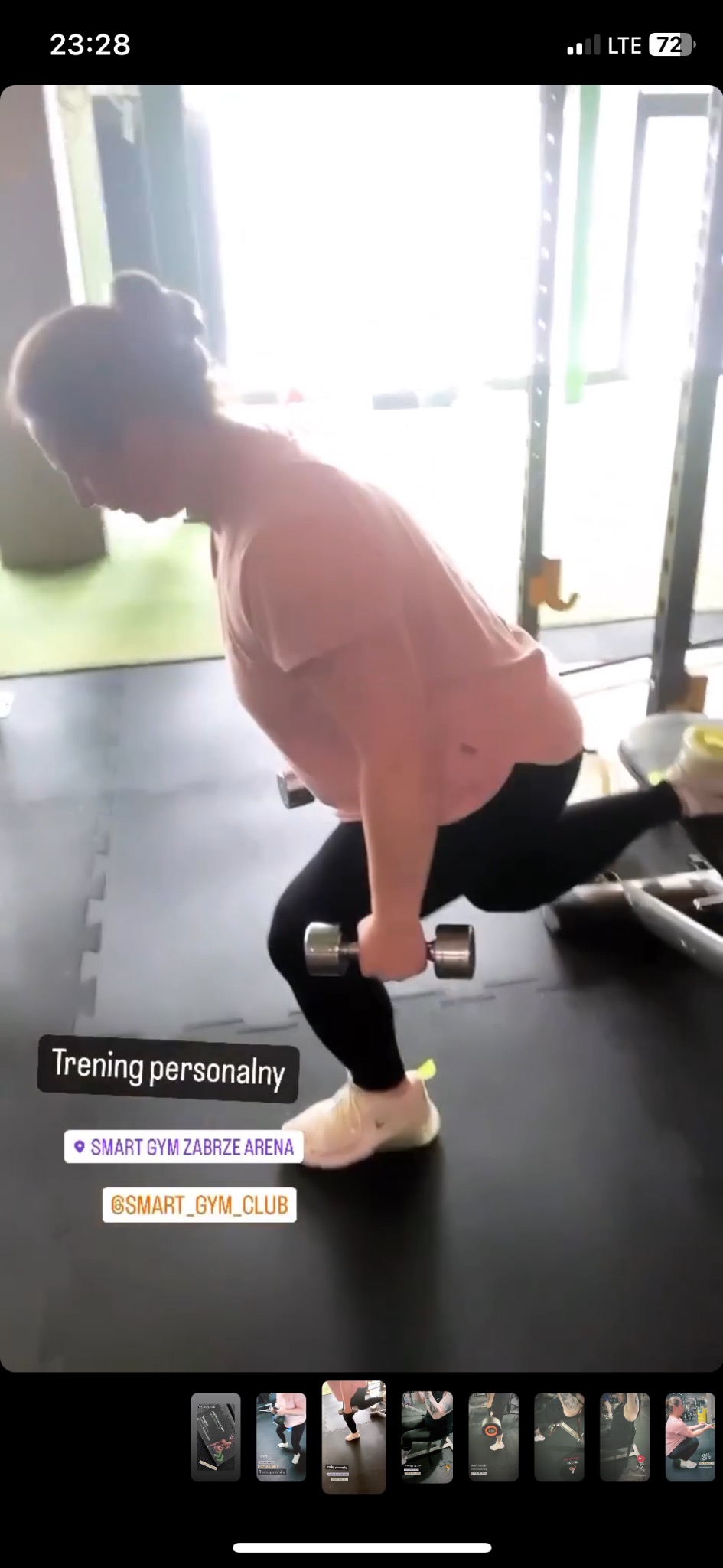 Kobieta w różowej koszulce i czarnych legginsach wykonuje wykroki z hantlami w siłowni, widoczna platforma do ćwiczeń i gumowa podłoga.