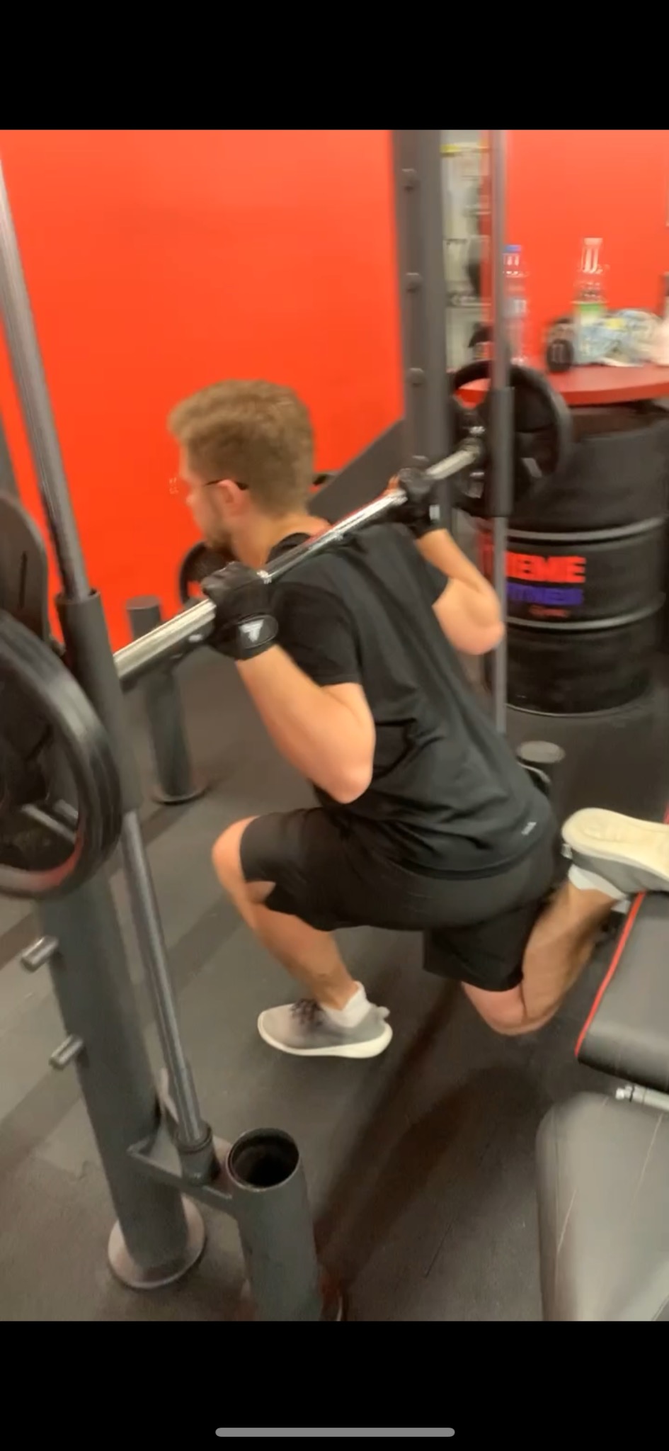Mężczyzna w czarnym stroju, wykonujący przysiad bułgarski ze sztangą w siłowni, wspierając tylną nogę na ławce, w tle widoczne butelki z wodą i pojemniki.