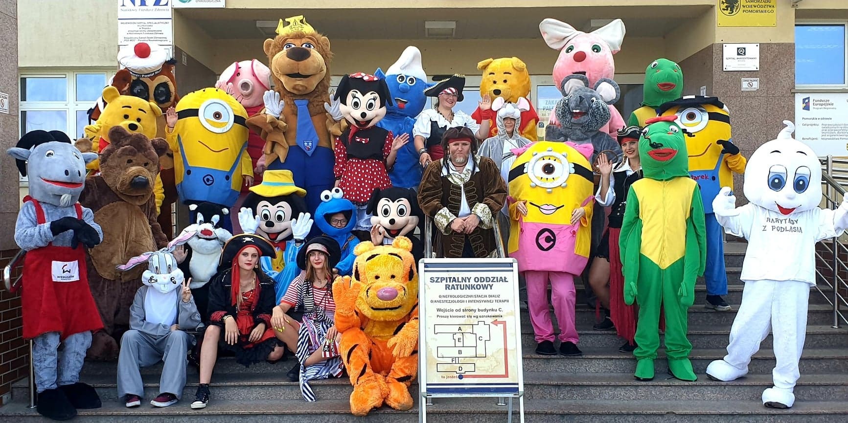 Grupa osób w kostiumach postaci z bajek i piratów pozuje na schodach przed budynkiem szpitala, obok tablicy informacyjnej z planem szpitala.