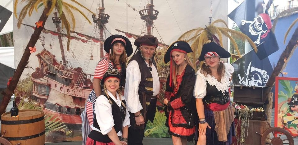 Grupa pięciu animatorów w kostiumach pirackich pozuje na tle dekoracji z motywem statku i palm, obok skrzyni ze skarbami i beczki.
